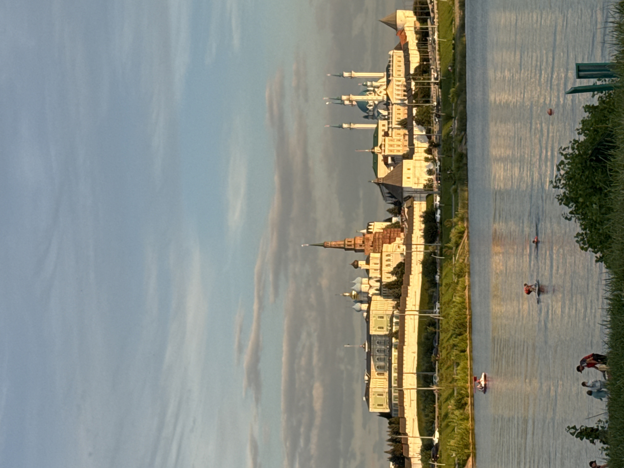 This is a wide shot of the Kazan Kremlin in Kazan, Russia, taken from across a body of water during the golden hour. The Kremlin, a UNESCO World Heritage site, sits atop a hill and is bathed in warm, late afternoon sunlight. Its iconic structures, including the Kul Sharif Mosque with its blue domes and minarets, and the Söyembikä Tower, are clearly visible. The white fortress walls of the Kremlin stretch across the horizon.
In the foreground, the water of the Kazanka River reflects the sky and the distant buildings. Several people are enjoying the water: two individuals are on a pedal boat, one person is paddleboarding, and a few others are swimming. On the grassy bank in the very front, a small group of people are gathered. The sky above is a soft blue with scattered clouds, tinged with the orange and yellow hues of sunset. Lush green trees and vegetation cover the slopes leading up to the Kremlin. The scene depicts a peaceful and picturesque urban landscape at dusk.