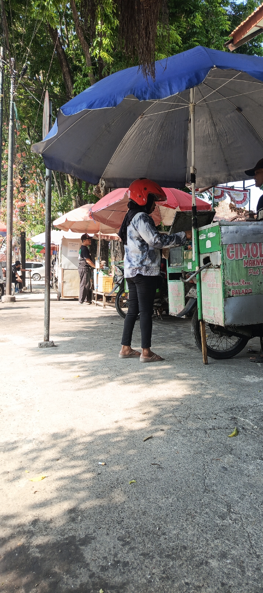 An outdoor scene in Krian, Indonesia, features two food carts operating under large umbrellas. The primary cart, made of green metal, displays branding that includes the word "CIMOL". A woman wearing a red helmet and a tie-dye jacket stands to the right of this cart, interacting with its contents. A man in a black shirt and pants stands behind a smaller, lighter-colored cart to the left, holding a container. In the background, trees and vegetation are visible, with dappled sunlight and shadows cast on the paved ground. The overall atmosphere suggests a bustling street food vendor area.