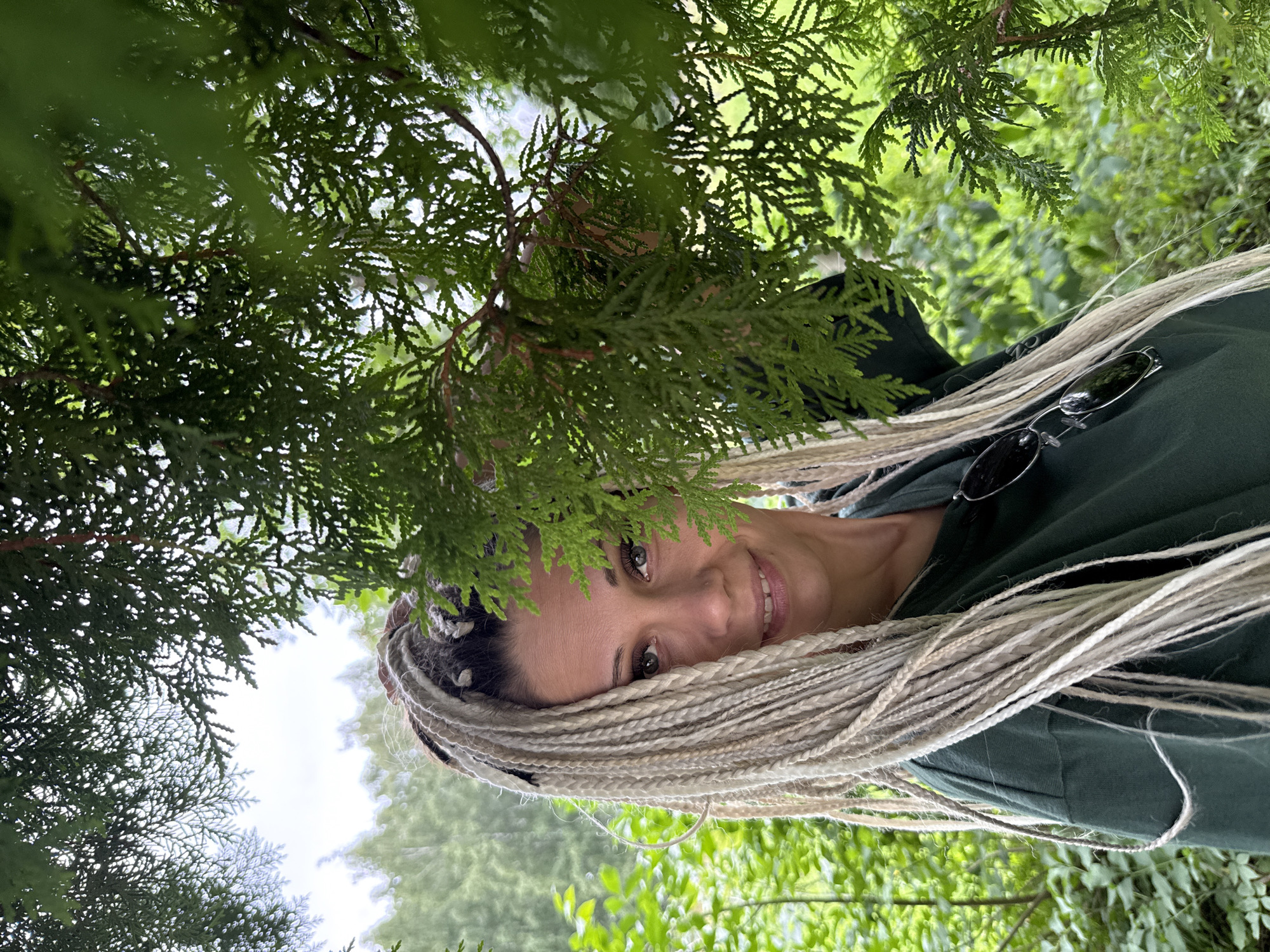 An outdoor, medium shot captures a woman with long, blonde dreadlocks looking directly at the camera. She is partially obscured by the branches of a dark green evergreen tree, with lush green foliage forming the foreground and background. The woman is wearing a dark green sweatshirt, and a pair of dark-lensed sunglasses are resting on the chest of her sweatshirt. Her expression is a subtle smile. The location is identified as Rusanovo, Russia.