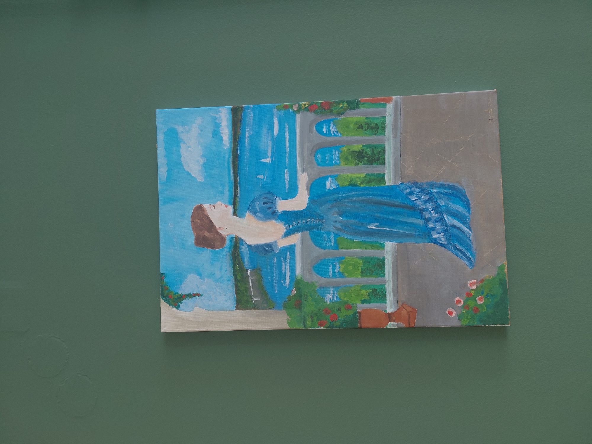 The image shows a painting of a woman in a blue dress on a balcony overlooking a body of water with sailboats. The painting is on a canvas and is hung on a dark green wall. The woman is standing with her back to the viewer, looking out at the water. She has her hair pulled up in a bun. The balcony has a white railing with arches. There are potted plants on the balcony. The body of water appears to be a lake or a bay. The sky is blue with some white clouds. There are trees and buildings in the distance on the far shore. The painting appears to be a realistic style. There is a signature in the bottom right corner of the painting that is illegible. There is no discernible location in the city of Municipal Unit of Aegio, Greece.