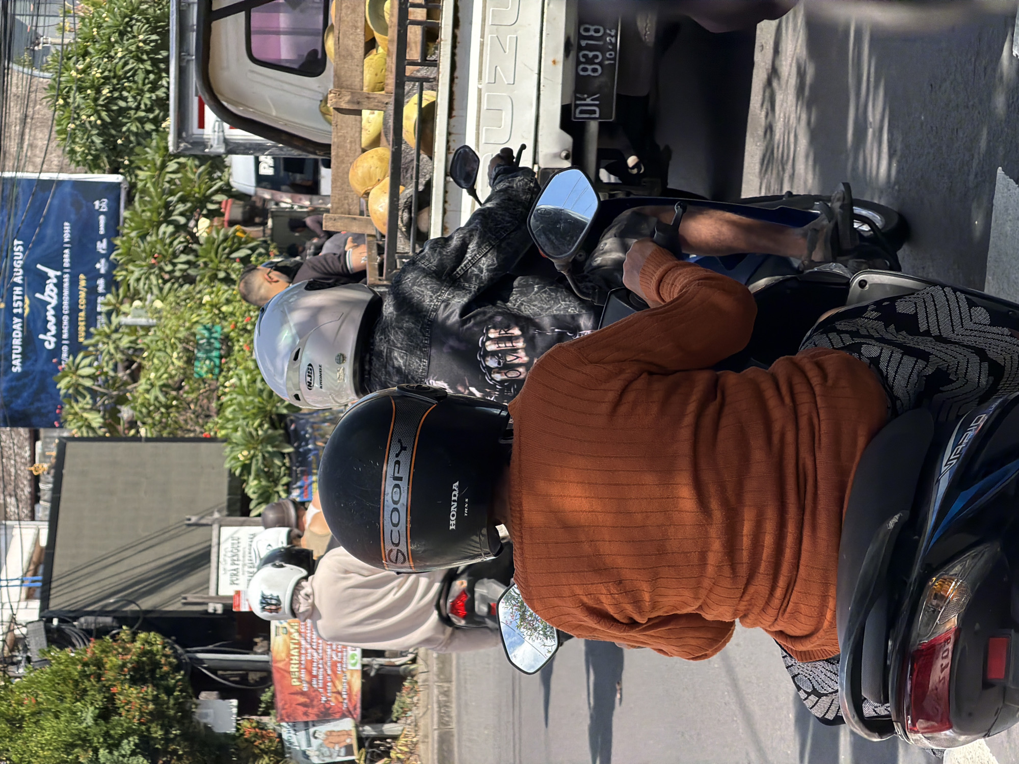 In North Kuta, Indonesia, on a sunny day, two individuals are riding motorcycles on a paved road. The foreground features a person wearing an orange knitted sweater and a black helmet with orange and silver accents and the text "SCOOPY" and "HONDA" visible. They are riding a black scooter, and the rear of the scooter is partially visible. Behind them, another person, also wearing a helmet, is riding a motorcycle. This individual is wearing a black jacket with a graphic design on the back that includes the word "OVER" in a bold font. A white truck, loaded with yellow fruits, is parked to the right. The truck has a license plate that reads "DK 8318 10.24". Buildings and lush green trees line the street in the background, suggesting an urban environment with significant vegetation. Billboards and signs are also visible, one of which has blue lettering that reads "SATURDAY 15TH AUGUST" and "chambord".