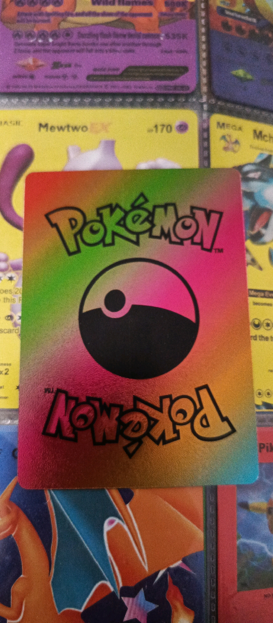 The image shows a collection of Pokémon cards. The foreground features a holographic Pokémon card with a rainbow-colored gradient. The Pokémon logo is visible in black, along with a stylized Poké Ball. The text "Pokémon" is also present upside down at the bottom of the card, also in black. In the background, other Pokémon cards are partially visible. One card appears to be yellow with text that reads "Mewtwo EX" and "-170". Another card is purple with text that is mostly illegible but appears to include "Wild Flames" and "Energy". A card with "MEGA" and "Mch" is visible to the right. The overall impression is that of a private collection of trading cards, possibly stored in a binder or album. There are no people or elements that would indicate a specific location in the city of Brwinów, Poland.