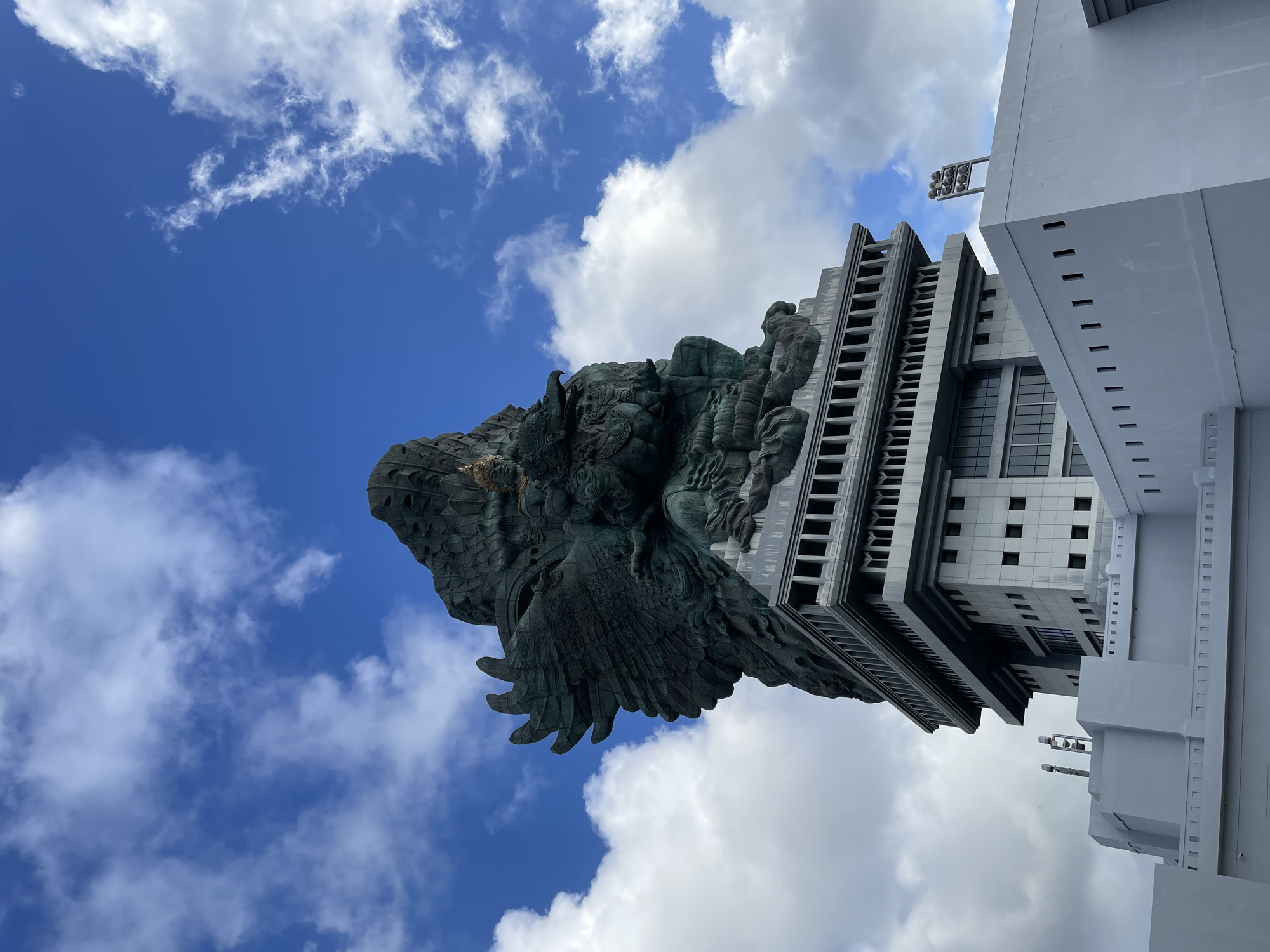 Captured from a low angle, the image showcases the Garuda Wisnu Kencana statue complex in Kuta Selatan, Indonesia. The bronze statue of Lord Vishnu riding the mythical bird Garuda dominates the frame against a backdrop of a bright blue sky dotted with puffy white clouds. Adjacent to the statue, a modern, multi-story white structure with a grid-like facade is visible. The perspective accentuates the statue's immense scale. The surrounding environment consists of buildings and sky, with no visible people.