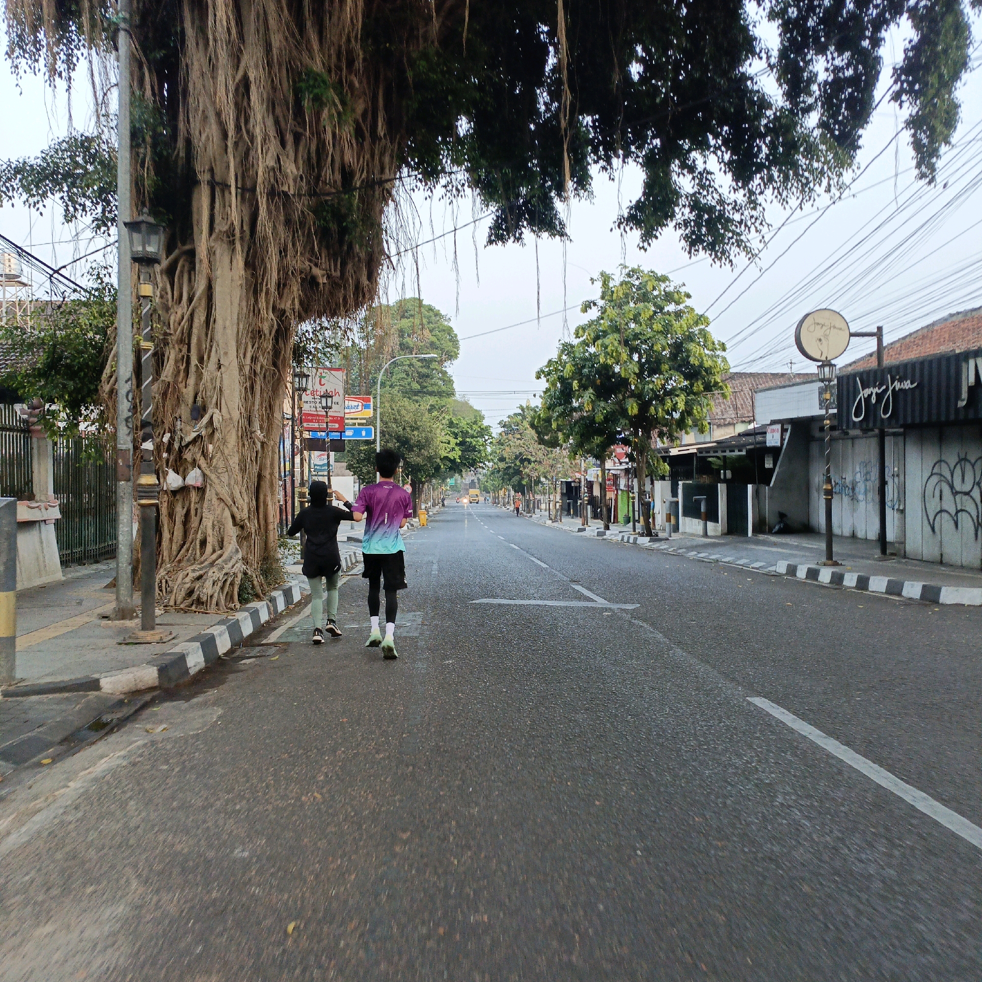 In Salatiga, Indonesia, an outdoor daytime scene unfolds on a street lined with trees and buildings. Two individuals, one wearing a black hijab and green leggings, and another in a purple and teal t-shirt and black shorts, are jogging away from the camera on the right side of the road. They are positioned on the asphalt, with the sidewalk to their left featuring a black and white striped curb and a large banyan tree with extensive aerial roots. A street lamp stands near the banyan tree. On the right side of the street, buildings with signs are visible, one of which reads "Joys Jiwa". Overhead, a tangle of electrical wires stretches across the sky. The overall atmosphere suggests an early morning or late afternoon activity, given the soft lighting.