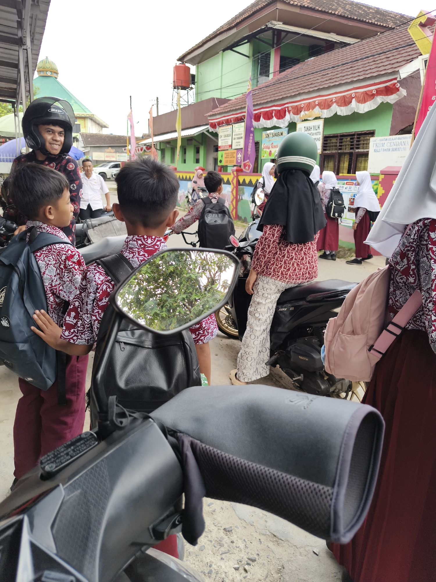 The image captures a street scene outside a school in Bandar Lampung, Indonesia, likely during school dismissal. In the foreground, a motorcycle is partially visible, with its handlebars and a side mirror reflecting a tree and sky. Behind the motorcycle, several children in school uniforms are gathered. Two boys in maroon and white patterned shirts and maroon pants with backpacks are standing. A girl wearing a black hijab, a white and maroon patterned top, and patterned pants is seated on the motorcycle. Another child in a white hijab and maroon uniform stands nearby. In the background, a man wearing a white shirt and a helmet is on a motorcycle, smiling at the children. Further back, a building with a green facade, red trim, and a tiled roof appears to be a school, with Indonesian signage visible, including what looks like "SMK SUKABUMI" and "FKM CENDANA". Red and white flags and banners decorate the school. The general environment appears to be a bustling urban setting with a mix of people and vehicles.