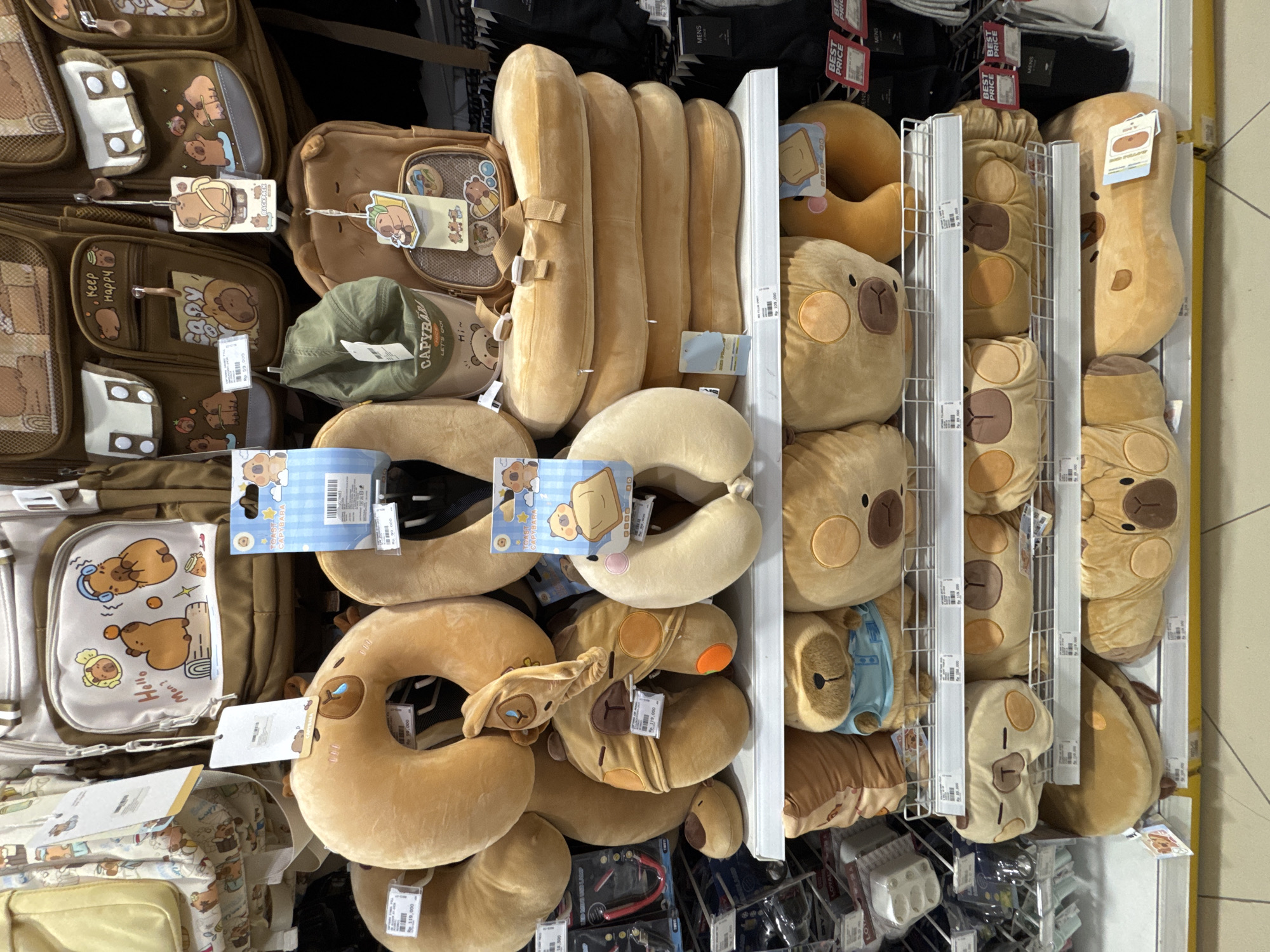 The image shows a retail display of merchandise in Kuta Selatan, Indonesia. The items primarily consist of capybara-themed products. The display includes backpacks, neck pillows, and plush toys arranged on shelves.