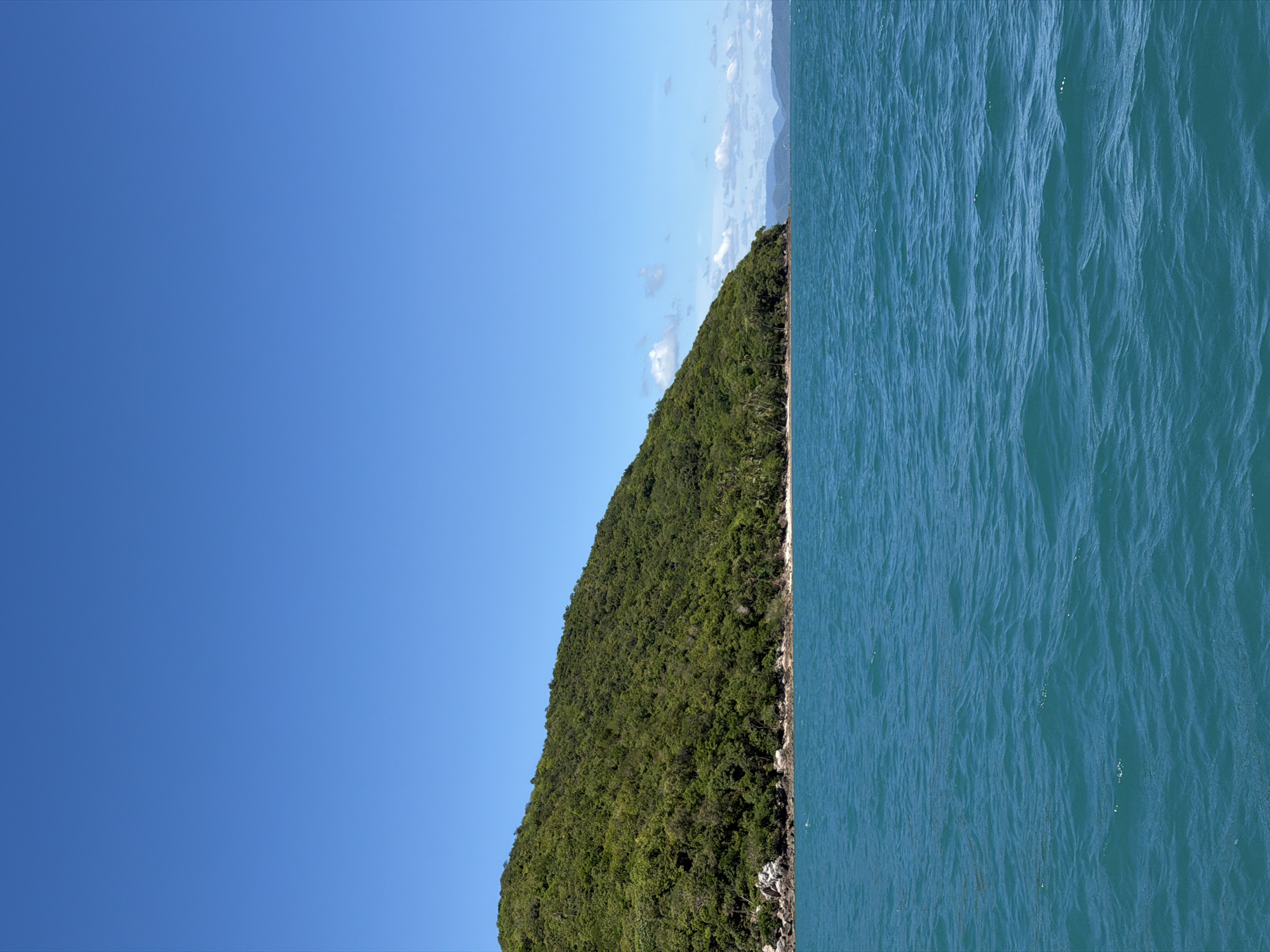 A vast, clear blue sky dominates the upper portion of the image. Below the sky, a lush, green, tree-covered hillside slopes down from the upper left to the middle right of the frame. The hillside meets a narrow strip of sandy beach. The foreground is filled with the tranquil, turquoise waters of the sea, showing gentle ripples and a few small whitecaps. The overall scene suggests a tropical island landscape, likely in Baan Thong Krut, Thailand, under a bright, sunny day. There are no people or animals visible in the image.