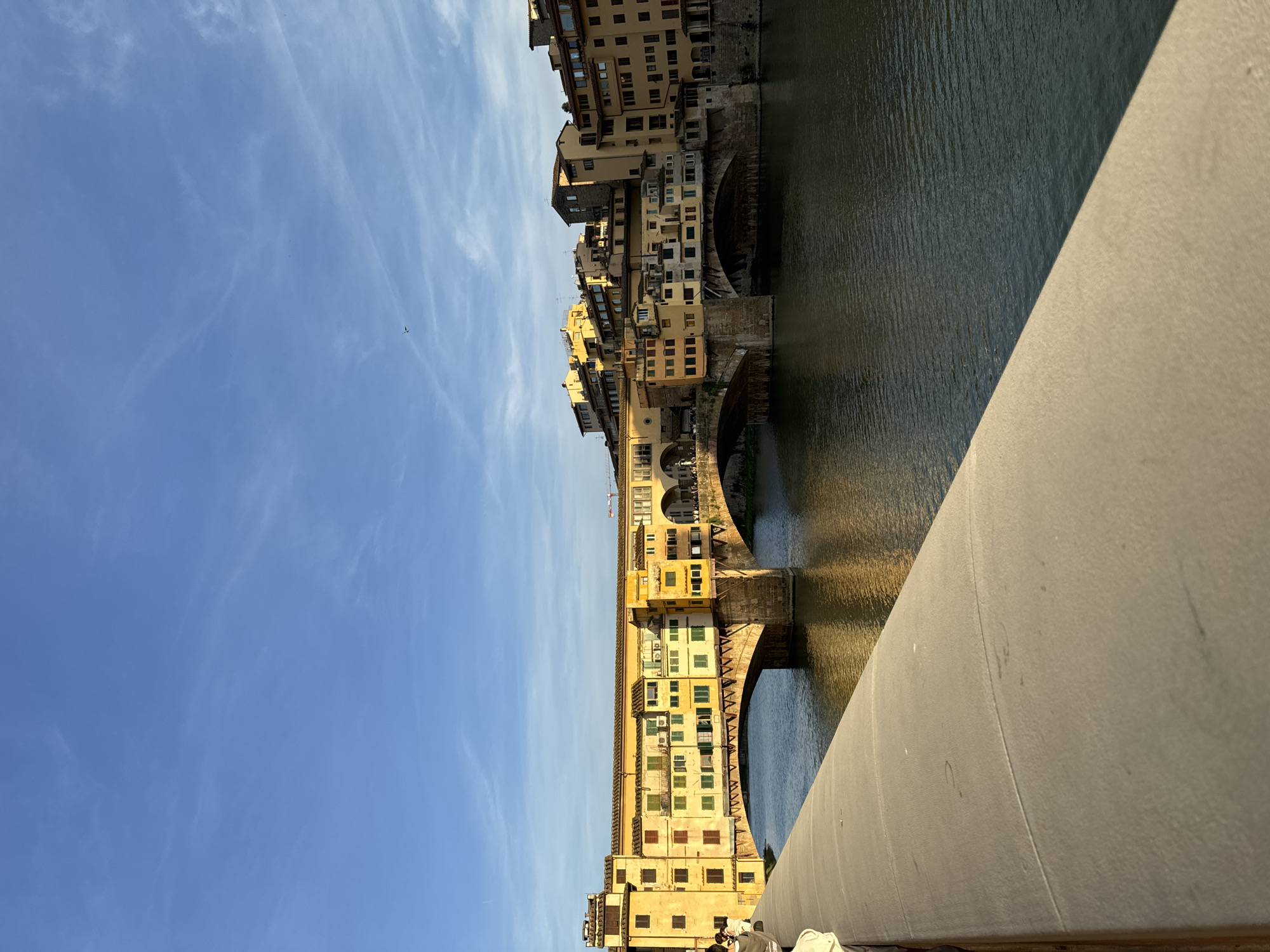The Ponte Vecchio, a medieval stone closed-spandrel segmental arch bridge over the Arno River in Florence, Italy, is depicted in this medium shot. The bridge is lined with shops built along it, a characteristic feature of this landmark. Buildings with multiple stories and various colored exteriors rise from the bridge's structure. The river, reflecting the buildings, flows beneath the arches of the bridge. A stone structure occupies the bottom right corner of the frame, possibly the edge of a walkway or a section of the bridge itself. Above the bridge, a clear blue sky is visible with a few wispy clouds.