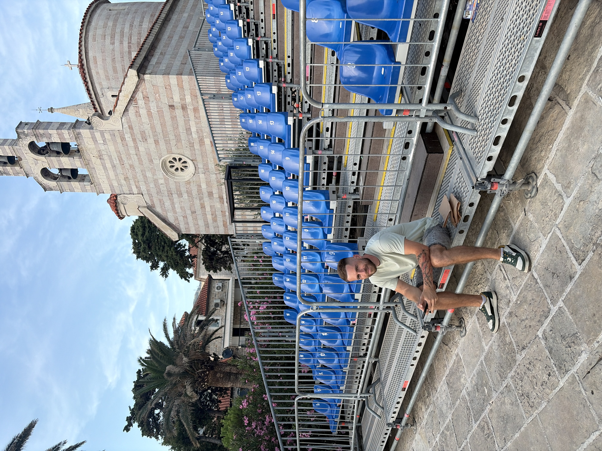 A man is sitting on a temporary seating platform in front of a church. The man is wearing a light green t-shirt, gray shorts, and green and white sneakers. He is holding a phone in his hands and looking towards the camera. Behind him, rows of blue stadium seats are set up, and beyond that, a stone church with a bell tower and red tiled roof is visible. Palm trees and pink flowering bushes are also in the background. The seating area and church appear to be located in the old town of Budva, Montenegro, likely near the Church of St. John the Baptist. The ground is paved with cobblestones.