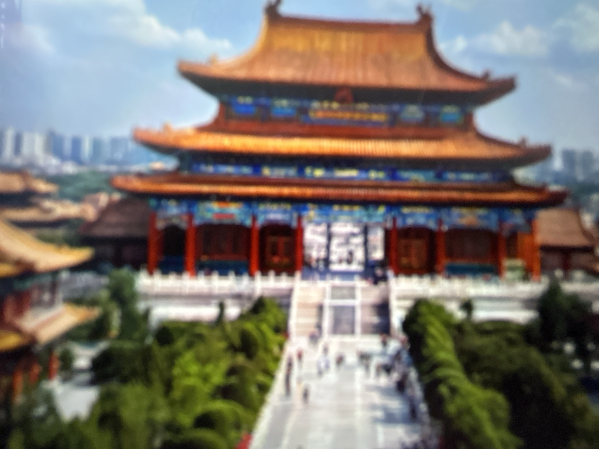 The image shows a blurred aerial view of a traditional Chinese-style building with a multi-tiered, ornate roof, possibly a temple or palace. The building is predominantly red and gold, with intricate decorative elements. To the left of the building is a large area of lush green trees and what appears to be a paved courtyard or plaza with some figures walking. In the background, modern high-rise buildings are visible, suggesting an urban setting. The sky is a light blue with some clouds. The image is oriented vertically and is of low resolution, making specific details difficult to discern. There is no indication of Gelendzhik, Russia, in this image.