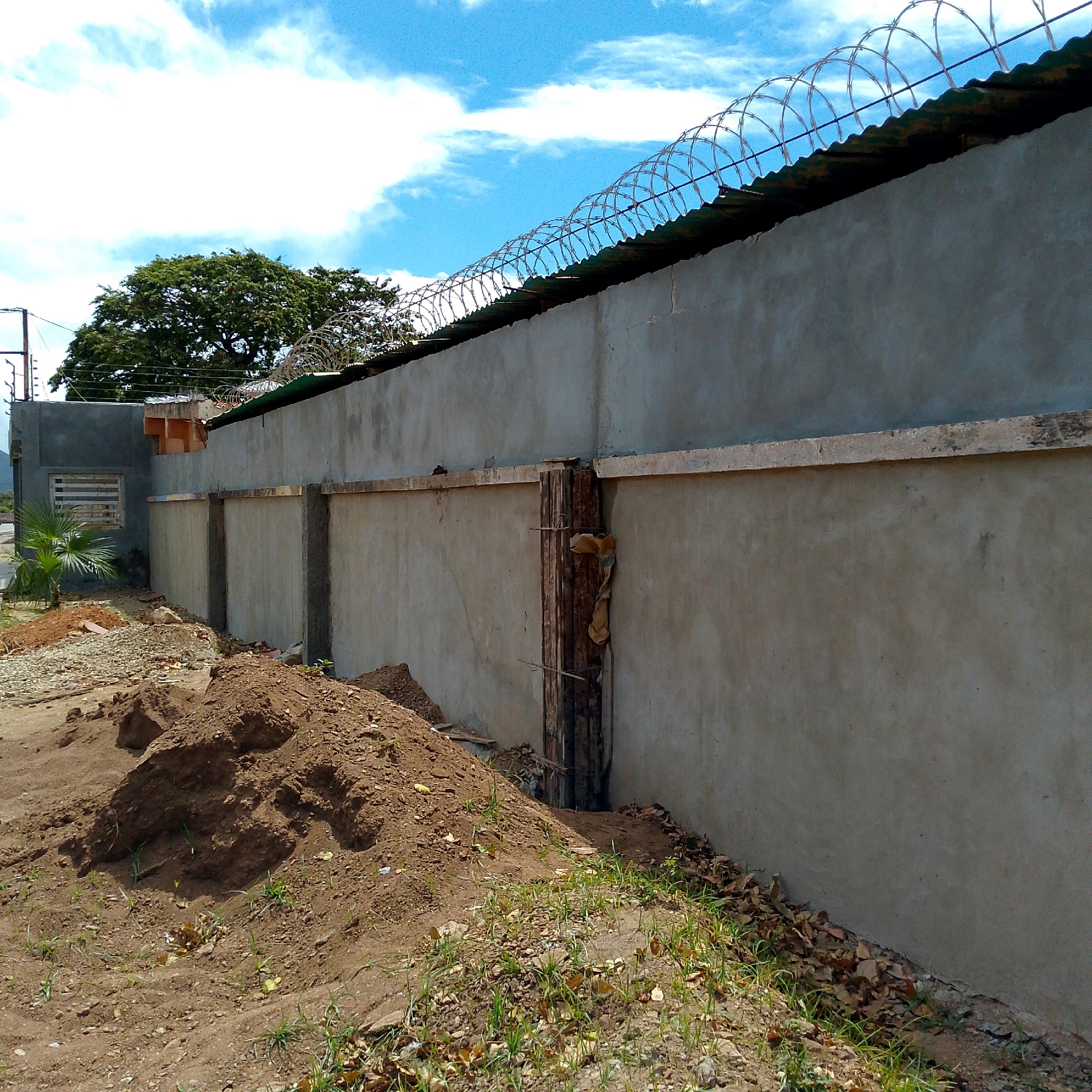 A daytime, medium shot depicts a tall, gray cement wall topped with razor wire in Altagracia, Venezuela. The wall runs horizontally across the frame, dividing the blue sky with scattered clouds from the ground. The ground consists of a dirt surface with patches of grass and fallen leaves. A pile of dirt is in the foreground. Vertical wooden supports are visible along the wall's length. A separate gray building with a barred window is to the left of the wall. A green tree is behind the wall.