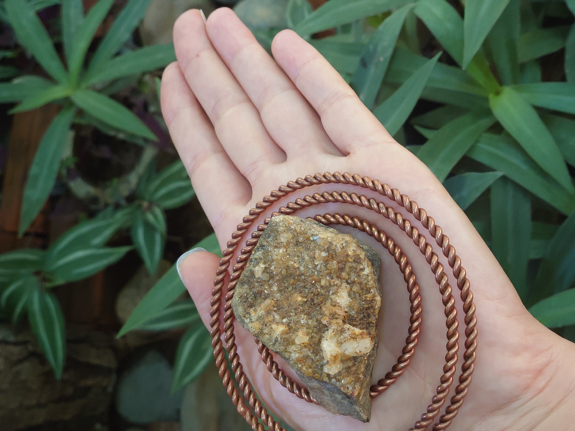 A hand holds a rough, tan and white rock fragment, possibly quartz or a similar mineral, nestled within three twisted copper wire loops. The rock is irregular in shape and appears to have a crystalline texture. The copper wire is a reddish-brown color and has a noticeable spiral pattern. The hand, which is fair-skinned, is open and cupped, with the fingertips and thumb visible. The background is blurred and consists of lush green foliage, with some darker, rough-textured elements, possibly wood or stone, in the upper left corner. The lighting suggests an outdoor, natural setting, possibly in a garden or natural landscape, with soft, diffused light. The scene is captured from a slightly elevated angle, looking down at the hand and rock.