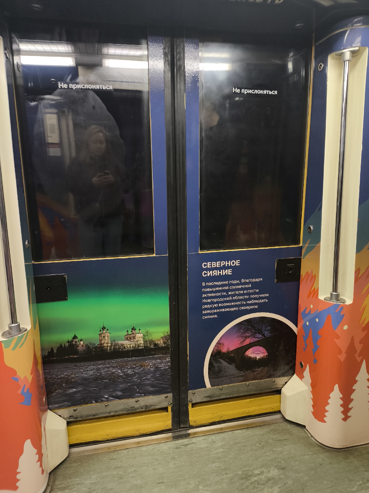 Here is a matter-of-fact description of the image:
The image shows a subway car door decorated with illustrations of the Northern Lights and Russian architecture. The upper section of the door has a window with the words "Не прислоняться" (Do not lean) printed on it in Russian. Below the windows, one door features an image of an aurora borealis over a cityscape, while the other features text explaining that increased solar activity has made the Northern Lights more visible in the Novgorod region, along with an image of a bridge at sunset. The sides of the doors are decorated with colorful, abstract patterns of trees. The flooring is a light green color. The location appears to be inside a subway car, possibly in Russia given the Russian text and architecture depicted.