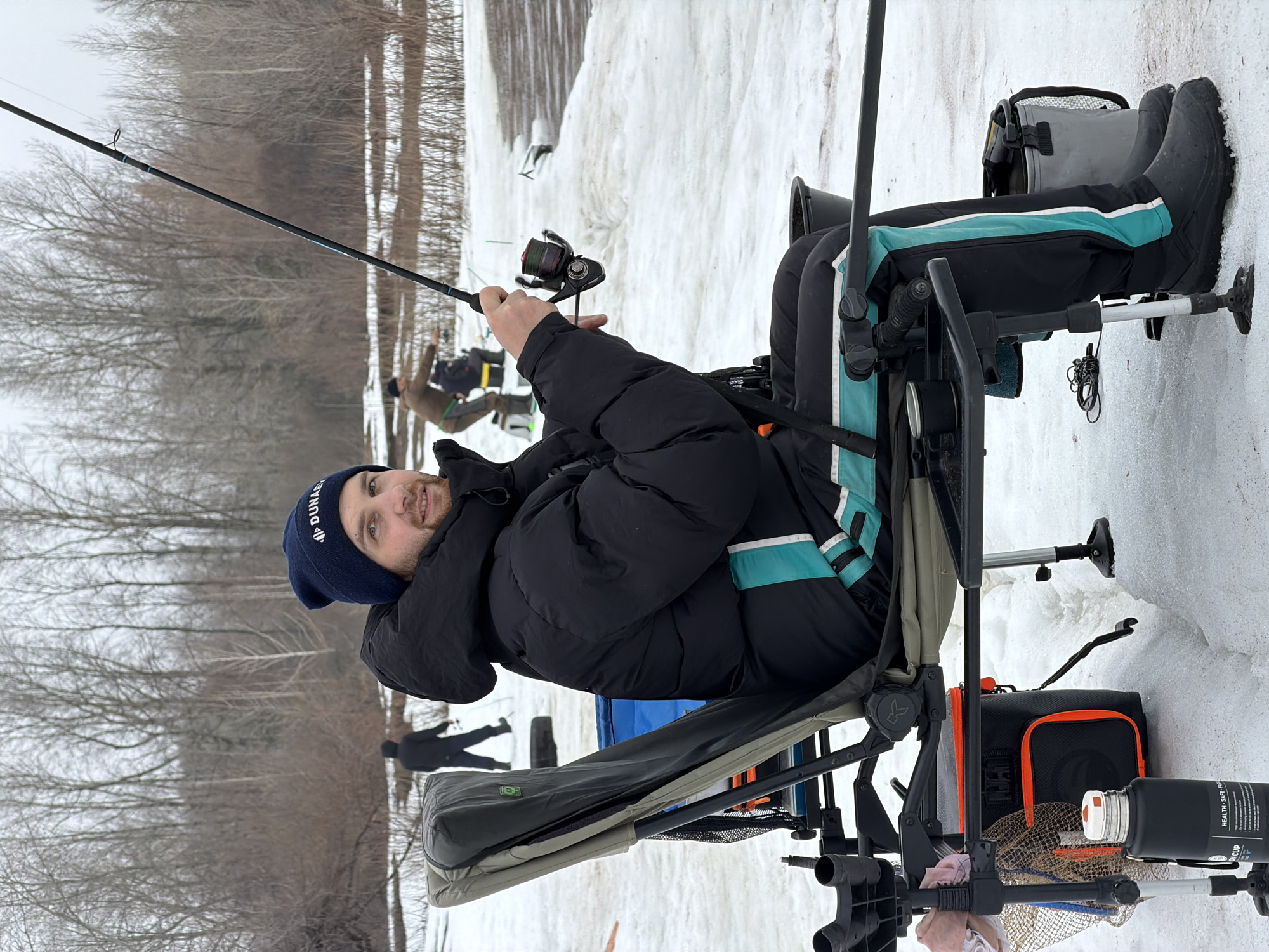 The image shows a man fishing in a snowy environment, presumably in the city of Chaikovsky, Russia. The man is seated in a specialized fishing chair, holding a fishing rod. He is wearing a black winter coat, dark trousers with a teal stripe, a dark blue beanie, and dark boots. The ground is covered in snow. Behind him, a group of bare trees is visible. There are other individuals visible in the background also fishing. Fishing equipment and accessories are attached to the chair.