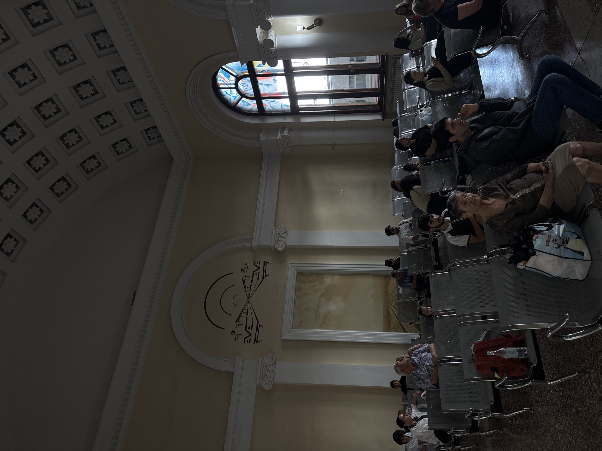 The image depicts the interior of a waiting area, possibly a train station, with people seated on rows of metallic chairs. The architecture features a high, decorative ceiling with a pattern of square panels and an arched window with stained glass. The walls are a light yellow color, and there is a large framed painting on the left wall depicting a landscape. Sunlight streams in from the stained-glass window on the right, illuminating parts of the room and casting shadows.
The people are diverse in age and gender, most are seated and appear to be waiting. Some are looking around, others are on their phones, and some are engaged in conversation. Several objects are visible on the chairs, including a red backpack with a water bottle, and bags.
Considering the architectural style, particularly the stained glass window and the decorative ceiling, and the presence of a painting that evokes a sense of travel or distance, this scene is likely from the Yerevan Railway Station, a significant transportation hub in Yerevan, Armenia. The station is known for its unique Soviet-era architecture, which often includes decorative elements and large windows.