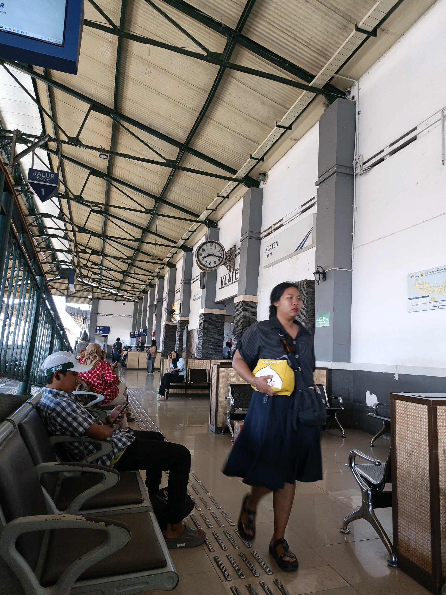 The image depicts the interior of a train station in Klaten, Indonesia, likely the Klaten Railway Station, given the signage. The scene is characterized by a high, corrugated metal roof supported by a dark green metal framework. White walls line the station, with some sections decorated with dark grey brickwork.
In the foreground, a woman in a dark blue dress walks from right to left, holding a bright yellow bag. Her footwear consists of dark sandals. To her left, a man sits on a bench, wearing a blue and white checkered shirt, black pants, and a white cap. He is looking at his phone, which he holds in his hand. His footwear consists of grey slip-on sandals. Behind him, other individuals are seated on similar benches, including a woman in a red and white patterned top and another woman wearing a hijab.
Various elements of the station environment are visible: a large clock with Roman numerals is mounted on a pillar, bearing the inscription "KLATEN". A sign indicating "JALUR TRACK 1" is also prominently displayed. Other details include benches for waiting passengers, lampposts, and a map posted on the wall. The floor is tiled with a pattern of raised ridges, likely for tactile guidance. The overall impression is one of a functional, somewhat weathered public transportation hub.