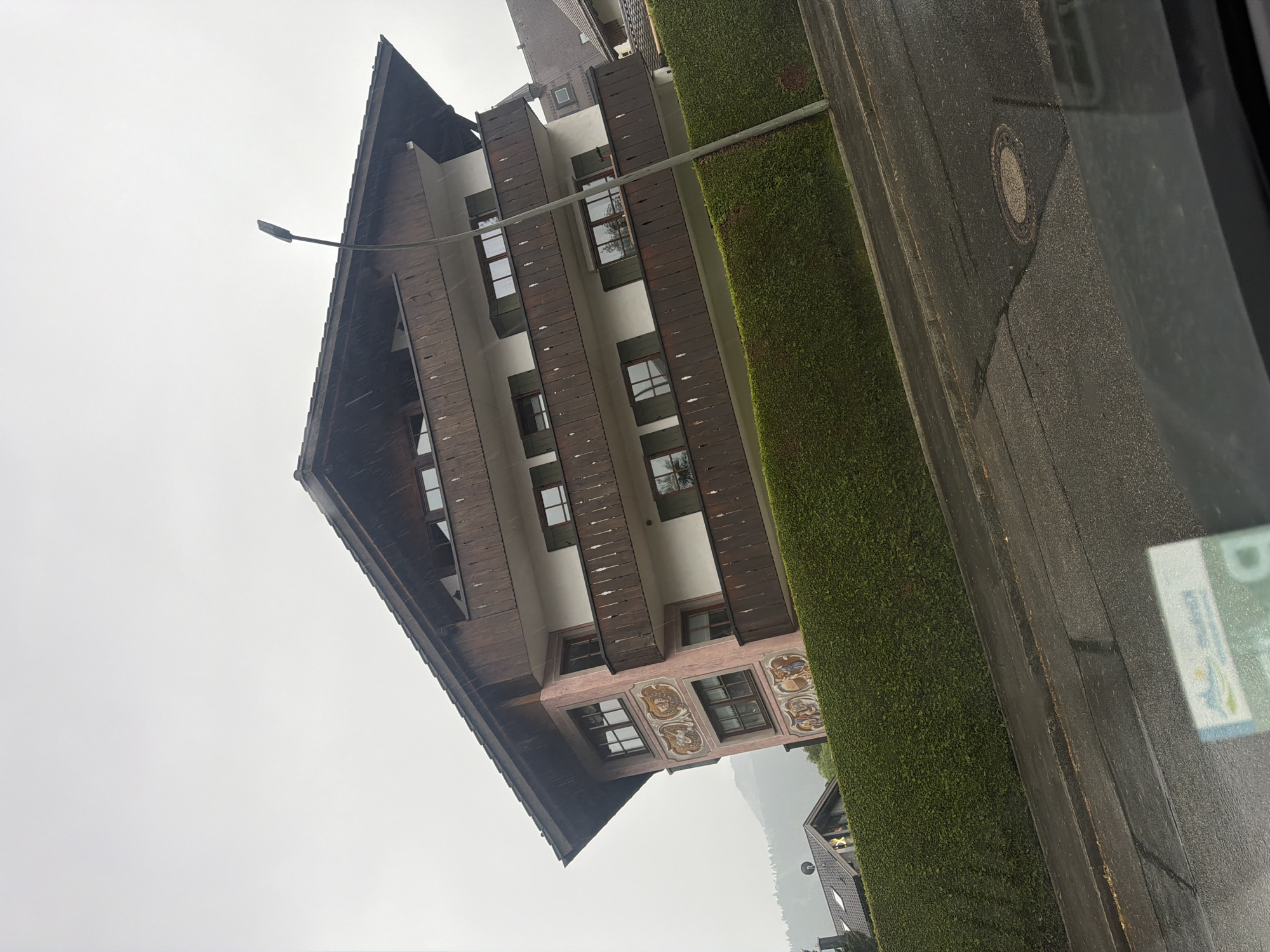 A building with a steeply pitched roof is viewed from the street. The building is multi-story, with wood detailing framing the windows and white walls. The lower level features painted ornamentation around the windows. To the right of the building is a tall, green hedge, and beyond that, an asphalt road. A manhole cover is visible in the road. In the distance, the hazy outline of mountains can be seen. A lamppost stands in front of the building. The scene appears to be viewed from inside a vehicle, due to the blurry edge on the right and a sticker on the window. The location is Unknown.