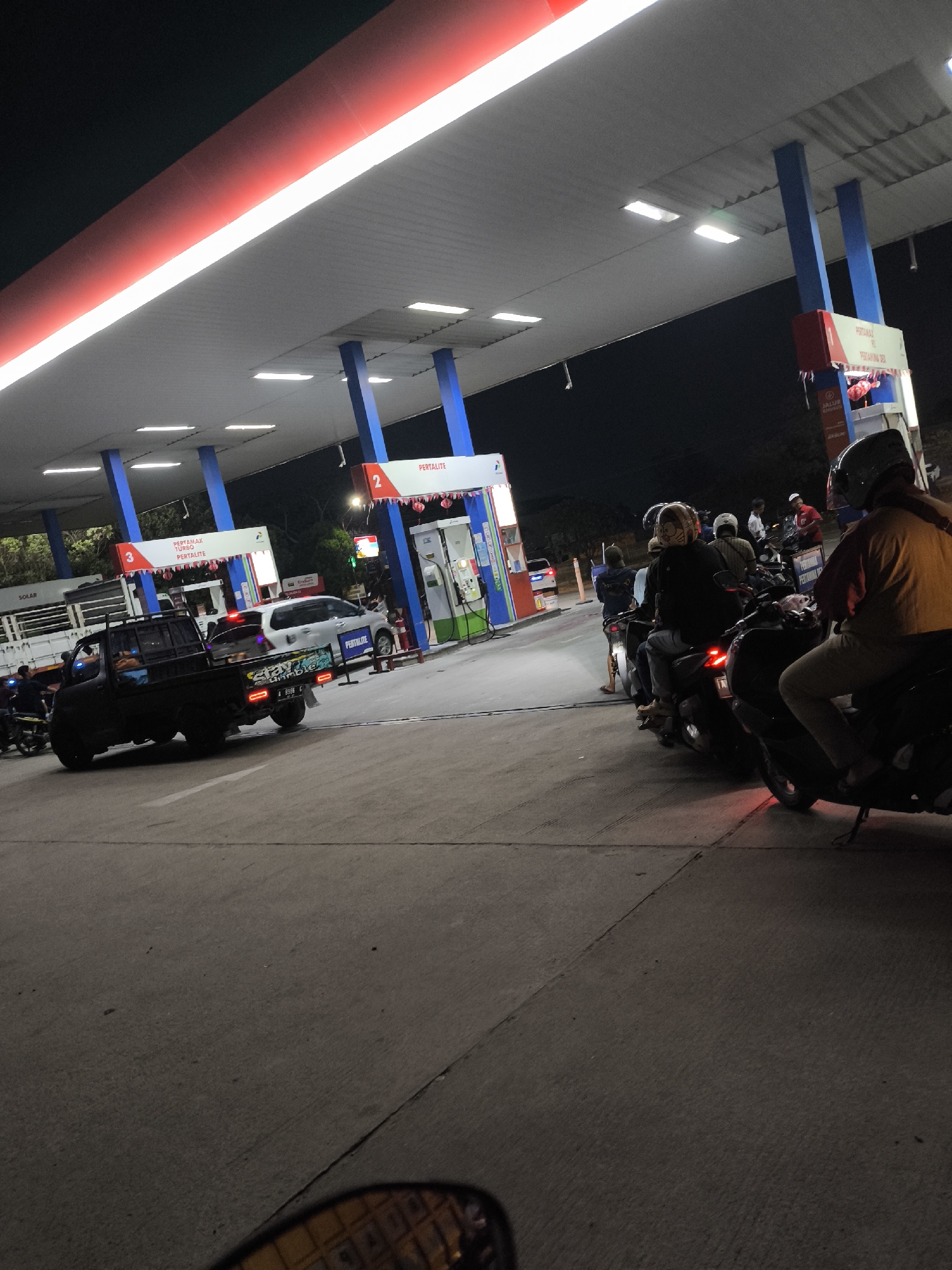 The image depicts a nighttime scene at a Pertamina gas station in Waringinkurung, Indonesia. A white van and a black pickup truck are visible, with several motorcycles lined up at the pumps. The gas station canopy is illuminated by red and white lights, and the sky is dark. A group of people, mostly on motorcycles, are waiting to refuel. The ground is paved with concrete, and there are visible lines and seams in the pavement. The overall atmosphere is that of a busy, functional service station at night.