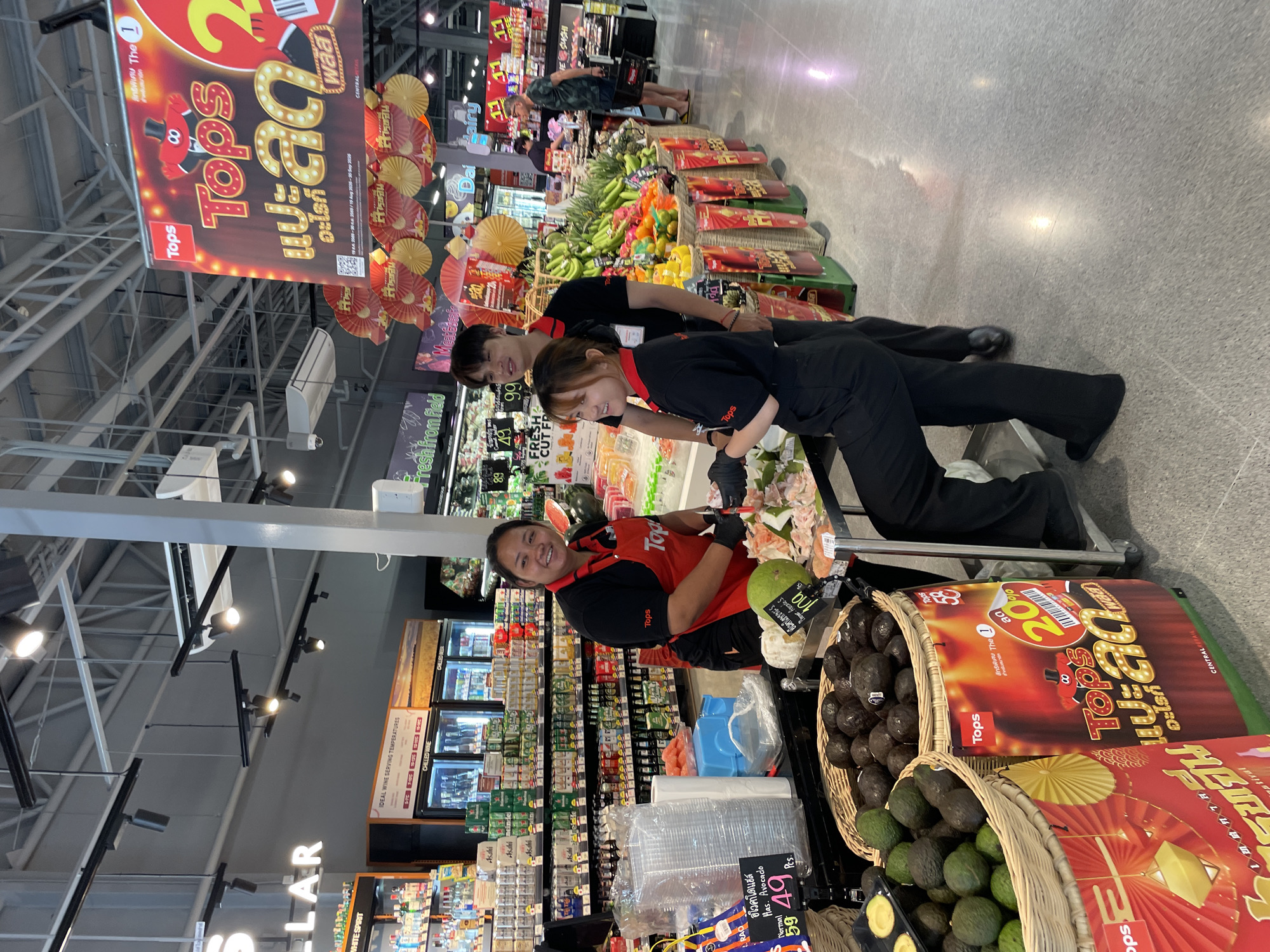 The image depicts an indoor grocery store environment, likely a supermarket, in Pattaya City, Thailand. In the foreground, a woman in a red apron and black uniform is standing behind a display of fresh produce, including avocados and what appear to be pomelos. She is smiling and holding a knife, suggesting she might be preparing or sampling the fruit. Two other individuals, a young man and woman, also in similar black uniforms with "Tops" logos, are standing nearby, possibly assisting customers or working. The shelves in the background are stocked with various food and beverage items, including bottled drinks and packaged goods. Hanging banners with promotional text and graphics are visible overhead, indicating a sale or special event. The floor is a polished gray material, and the lighting is bright and artificial, typical of a retail setting. The overall scene captures a moment of commercial activity within a well-lit supermarket.
