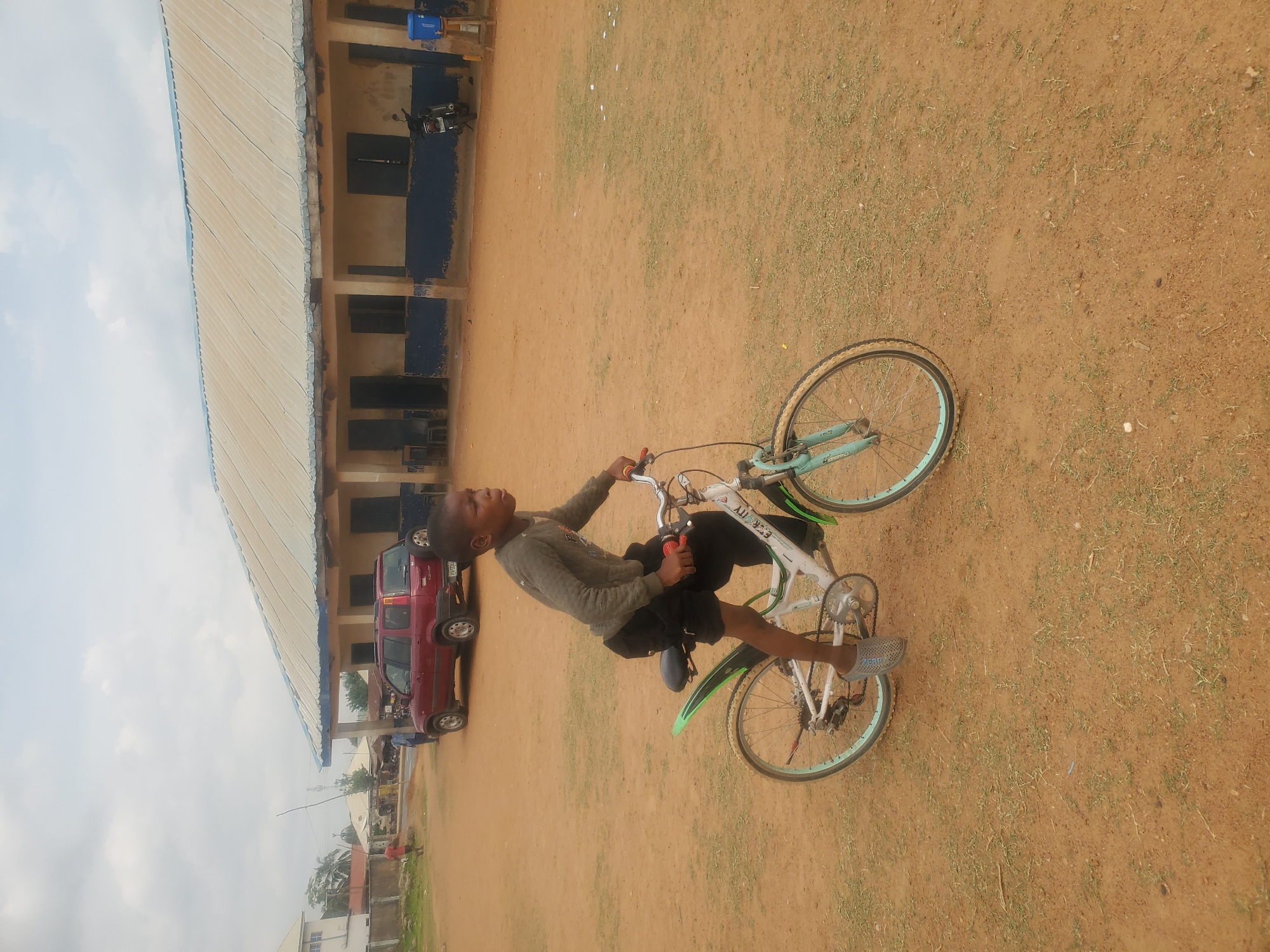This outdoor, portrait-oriented image, taken in Akwanga, Nigeria, captures a young boy sitting on a bicycle on a light brown dirt ground. The boy, who appears to be of school age, is dressed in a grey long-sleeved shirt and dark shorts, with light grey slip-on shoes. He is positioned on a light blue and white bicycle with green mudguards. The text "A.U.R.E.S.S." is visible on the bicycle's frame. The boy is looking upwards and slightly to his left, with a neutral expression.
The setting is an open area dominated by light brown soil with scattered patches of green grass. In the background, to the left of the boy, is a single-story building with cream-colored walls and a corrugated metal roof. The building features blue-painted lower sections on its pillars and walls. Several dark, possibly glass, window openings are visible. Parked next to the building is a red car, appearing to be an SUV or wagon, and a motorcycle is also visible further along the building. The sky is bright with light clouds, indicating daytime. The overall lighting suggests a clear day. The scene appears to be a casual moment in a semi-urban or rural environment.