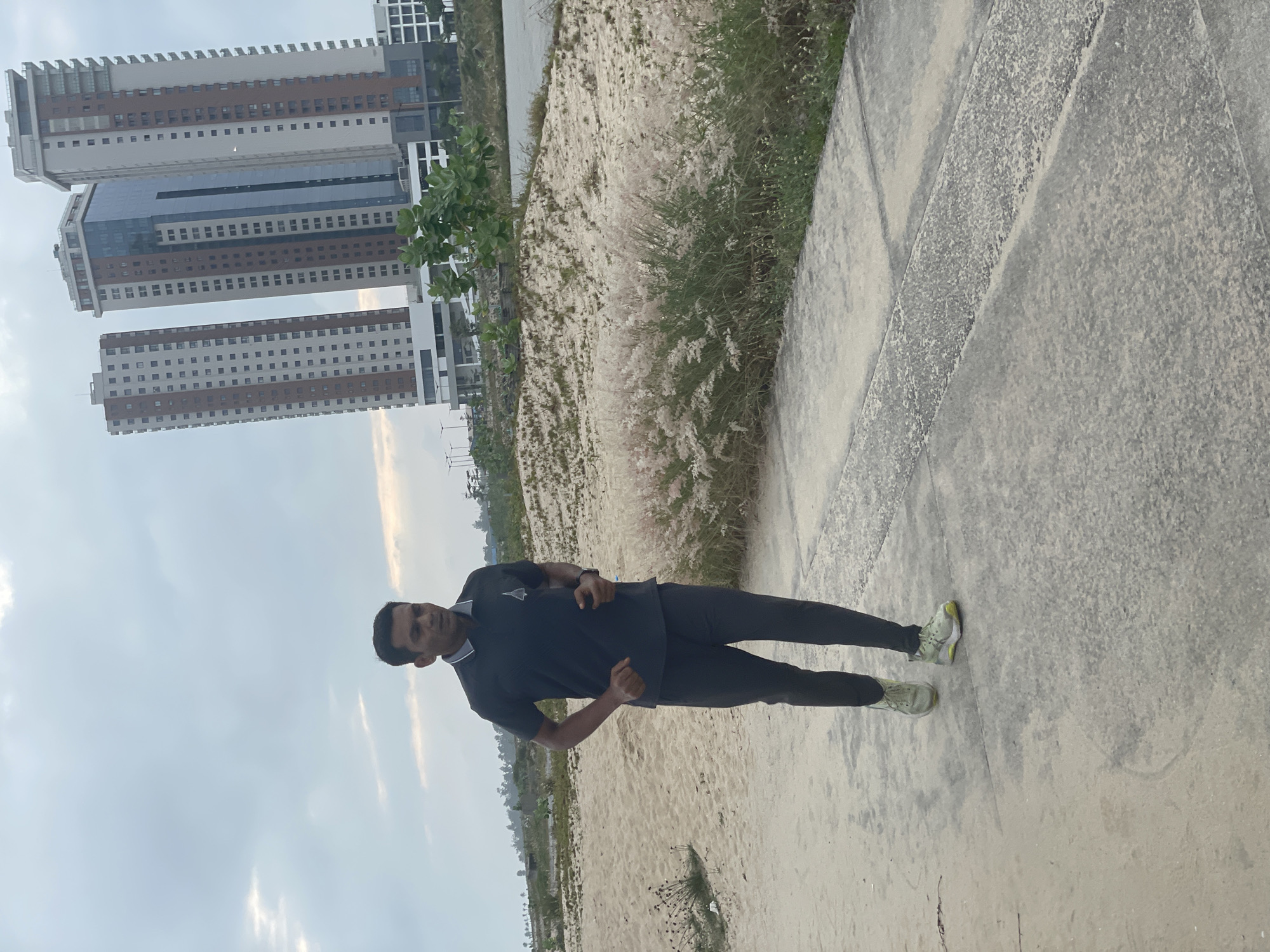 A man of Indian descent is captured mid-stride while jogging on a paved path. He is wearing a dark polo shirt with a grey collar and dark trousers. His athletic shoes are light-colored with yellow accents. The man is positioned on the left side of the frame, facing towards the right. In the background, a sandy embankment is dotted with dry, wispy grass. Beyond the embankment, a body of water is visible, and further in the distance, three tall, modern buildings rise against a cloudy sky. These buildings are located in the city of Inupa Kekere, Nigeria. The overall scene suggests an outdoor recreational activity in a developing urban environment.