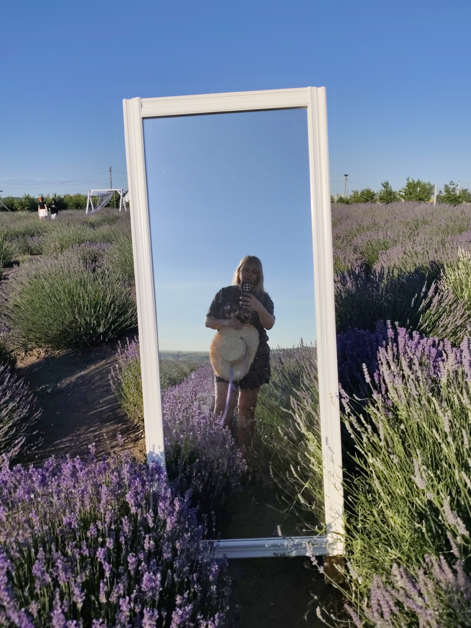 In Nechaiane, Ukraine, a woman stands in a lavender field, holding a straw hat and a smartphone, taking a selfie in a freestanding mirror. The mirror reflects the woman, the lavender field stretching behind her, and a clear blue sky. The field is densely planted with rows of blooming lavender bushes. In the distance, two other figures are visible, as well as a white swing set. The scene is illuminated by bright sunlight, casting shadows on the ground.