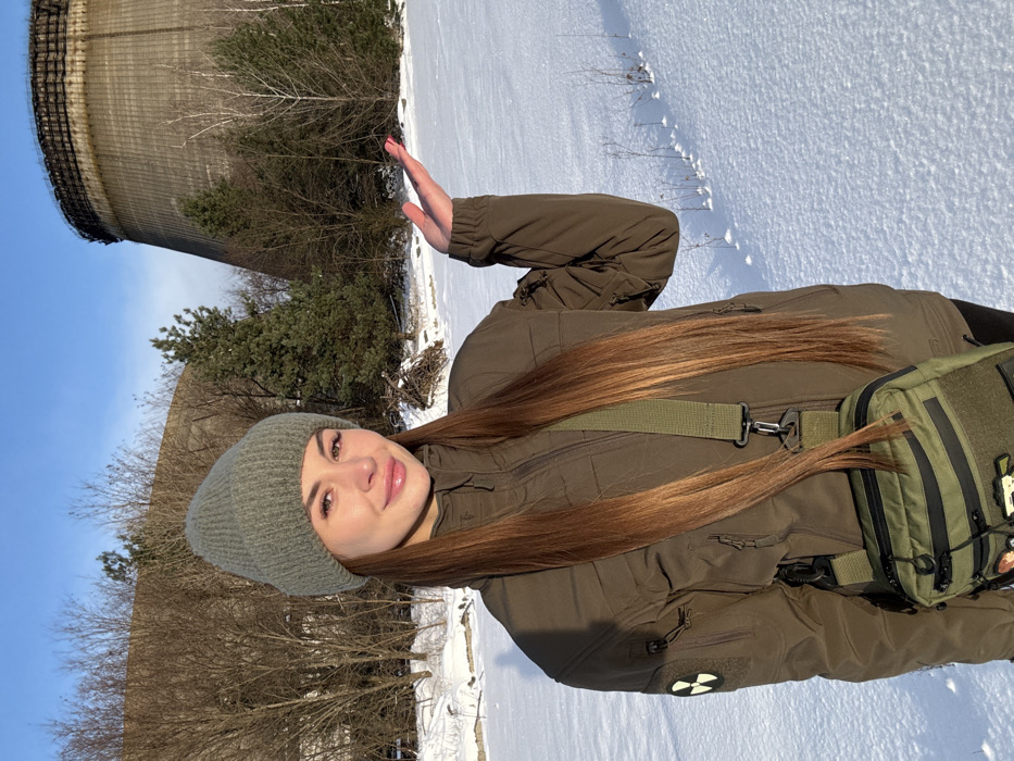 A young woman stands outdoors in a snowy winter landscape, with a prominent derelict cooling tower in the background, strongly suggesting a location like Pripyat, Ukraine, reinforced by the radioactive symbol patch on her jacket. She has long brown hair and is dressed in winter attire: a grey knit beanie, an olive green puffer jacket, and a matching olive green shoulder bag. She smiles towards the camera, her right hand raised in a wave.
The setting is bright and sunny with a clear blue sky. Bare deciduous trees and evergreen pines dot the snow-covered ground. The large, concrete, derelict cooling tower dominates the left background, appearing weathered. A yellow circular patch with a black radiation hazard symbol is visible on her left jacket sleeve. The ground is uniformly covered in fresh white snow. No explicit text is visible in the image beyond the symbol.