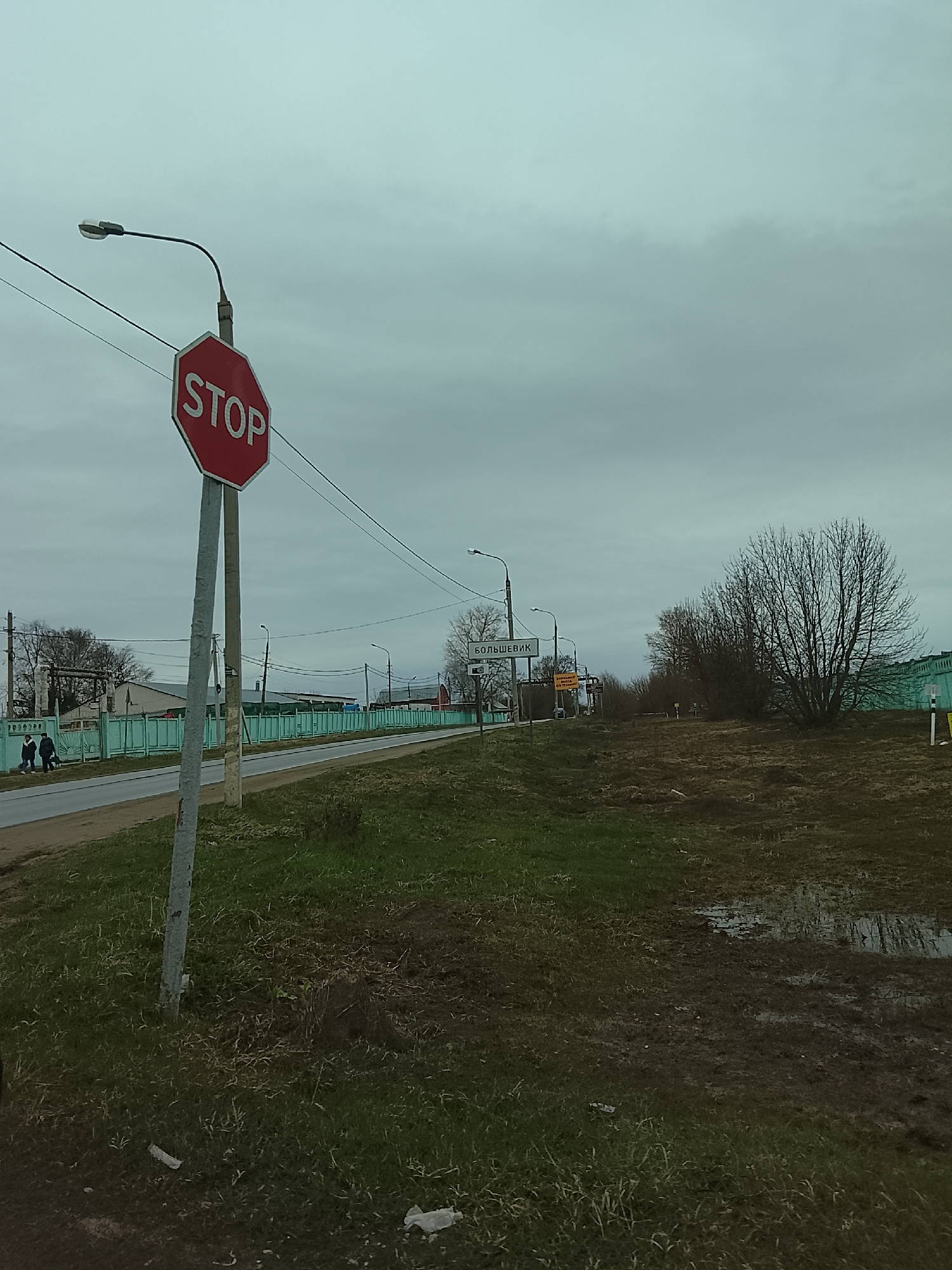 The image depicts a scene in Dashkovka, Russia. A stop sign is prominently displayed next to a road. Further down the road is a sign that reads "Большевик," indicating the location of the Bolshevik settlement. Two people are walking on the side of the road. The environment includes overcast skies, a road with a green fence, grassy and muddy terrain with puddles. Bare trees are visible in the background. Street lamps are present along the road.