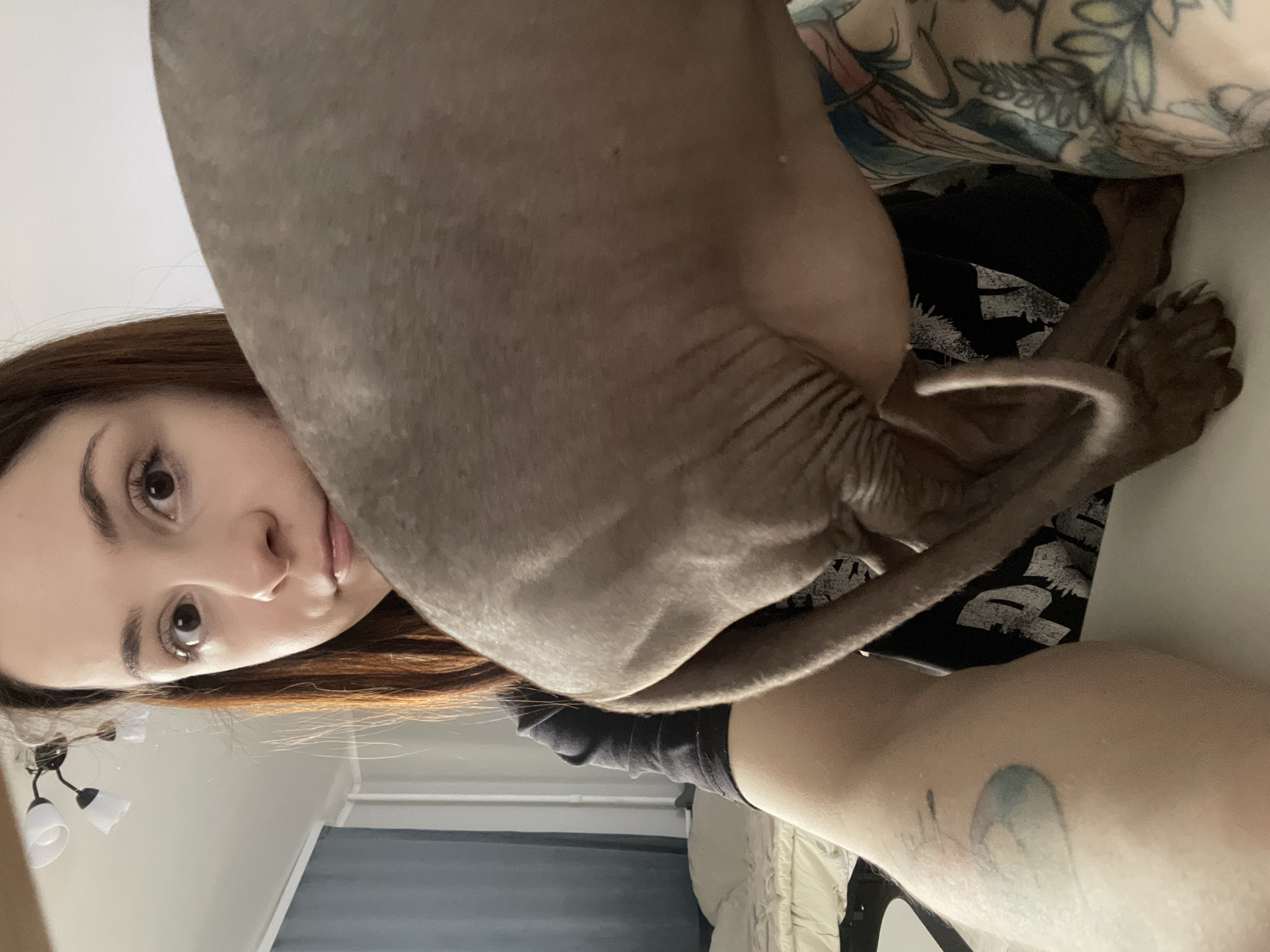 The image is oriented sideways, with the top of the scene on the left and the bottom on the right. A young woman with reddish-brown hair is partially visible on the left side of the frame, her face angled towards the camera. Her right eye, eyebrow, nose, and part of her mouth are visible, conveying a neutral or slightly inquisitive expression.
A Sphynx cat, with greyish-brown, wrinkled skin, dominates the central and upper-right portions of the frame, positioned very close to the camera. Its back, hindquarters, and long, thin tail are prominent, along with one hind paw displaying noticeable claws. The cat appears to be resting on or near the woman.
The woman has several tattoos: a large, colorful design featuring abstract or floral elements is visible on her upper arm or shoulder area, and a smaller, lighter, possibly blue-toned tattoo is on her lower arm or leg. She is wearing a dark garment.
The background on the far left reveals a white wall or ceiling. Below the woman's head, a light fixture with two white, inverted-cup shades is present. Further towards the bottom right, a dark window blind or curtain is visible against a wall, next to light-colored bedding, indicating an indoor setting. The overall lighting is bright, suggesting daytime. The image captures a close-up, intimate moment between the woman and her pet. There is no visible text.