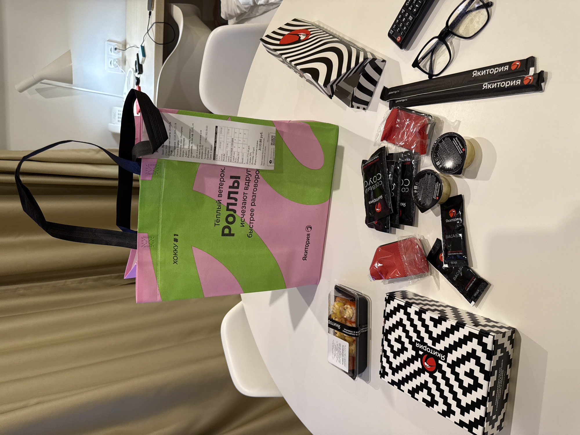 Here's a detailed description of the image:
The image shows a white, round table cluttered with food items and accessories from the restaurant "Yakitoriya". The items are likely a takeout or delivery order.
Prominently featured is a reusable shopping bag with pink and green sections. Text on the bag in Russian includes phrases related to the restaurant and suggests the items are "rolls", "vanish", and "faster conversation." A receipt is partially visible on the bag, mentioning "Khokku #1".
Next to the bag, several takeout containers and condiment packets are scattered. These include sealed containers with what appears to be soy sauce, wasabi, and ginger. Two sets of chopsticks are sealed in black paper marked with the Yakitoriya logo. There are also two containers of sushi.
Other items on the table include a remote control, a pair of glasses, and two boxes. The boxes have distinctive black and white zebra-like and zigzag patterned designs.
The text on the receipt includes Russian place names and addresses and specifies that the order was placed at a Yakitoriya restaurant near the Technopark metro station in Moscow, Russia, at Andropov Avenue 10.