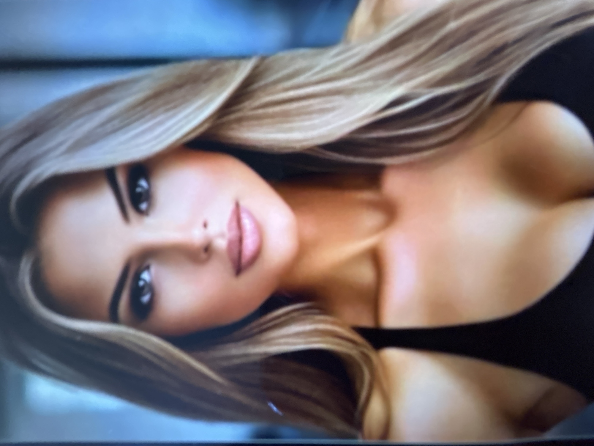 This media file features a close-up portrait of a person with blonde hair. The subject is positioned on their side, with their face turned towards the viewer. Their hair is long and styled in loose waves, framing their face. The subject has fair skin, dark eyebrows, and appears to be wearing makeup. Their eyes are looking directly at the camera, and their lips are slightly parted, revealing a soft pink color. The image is a close-up, focusing on the upper body and face, with a black garment visible around the chest area. The background is blurred and appears to be a light blue or grey color, suggesting an indoor or studio setting. No specific activity or event is depicted, and there are no discernible time of day, weather, or location cues beyond the provided context of Gelendzhik, Russia. There is no visible text in the image.