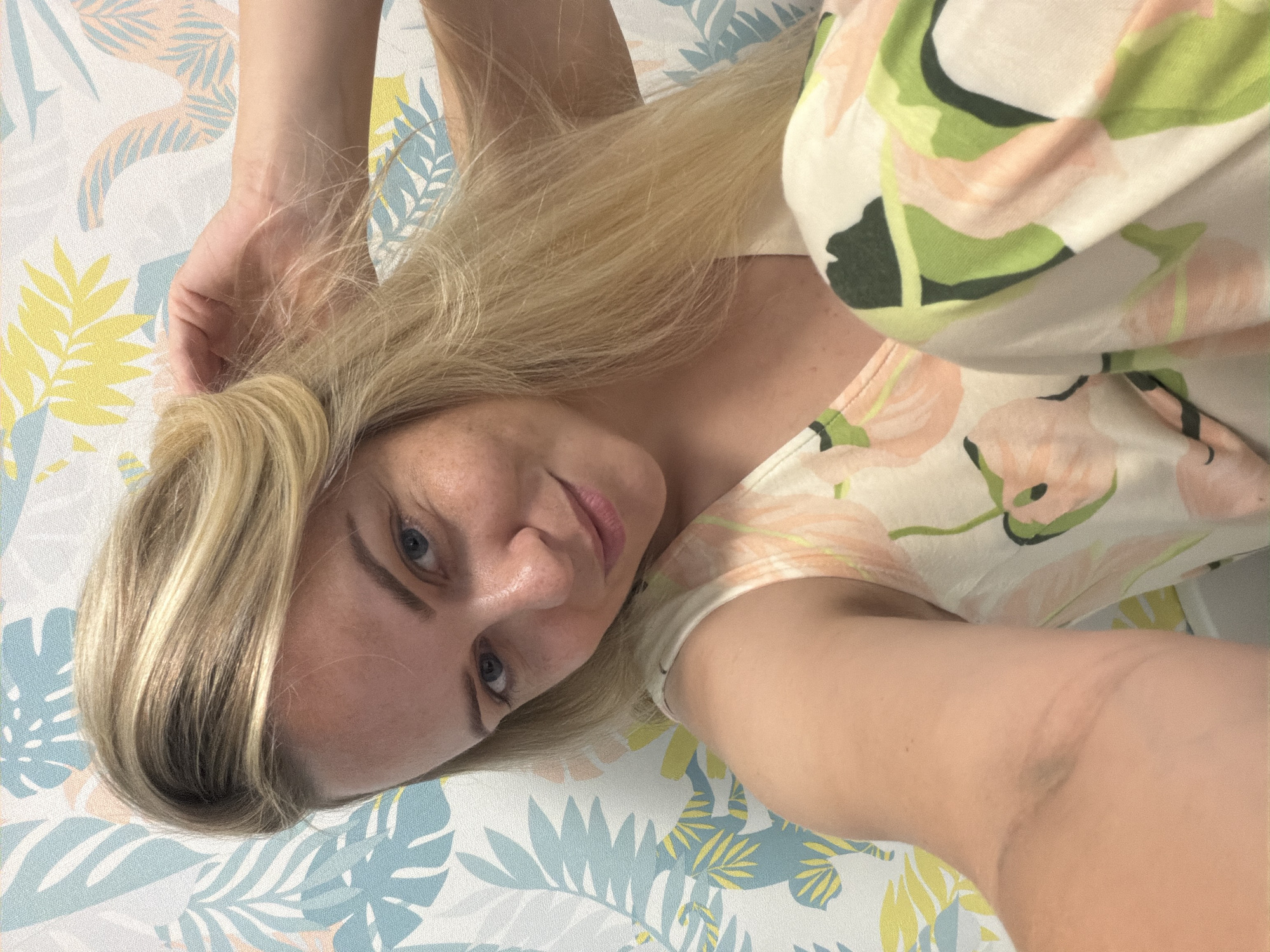 The media file is a close-up, vertically oriented selfie of a young woman with blonde hair. She is lying down on a surface with a floral and leaf pattern in shades of blue, yellow, pink, and green. She is wearing a light-colored t-shirt with a similar floral print. Her left arm is raised and bent, with her hand touching her hair near the back of her head. Her face is turned towards the camera, with her eyes looking slightly off to the side. Her expression appears relaxed and content. The lighting suggests it is daytime, possibly indoors given the texture of the patterned surface which could be wallpaper or fabric. No text is visible in the image. The location context is Votkinsk, Russia.