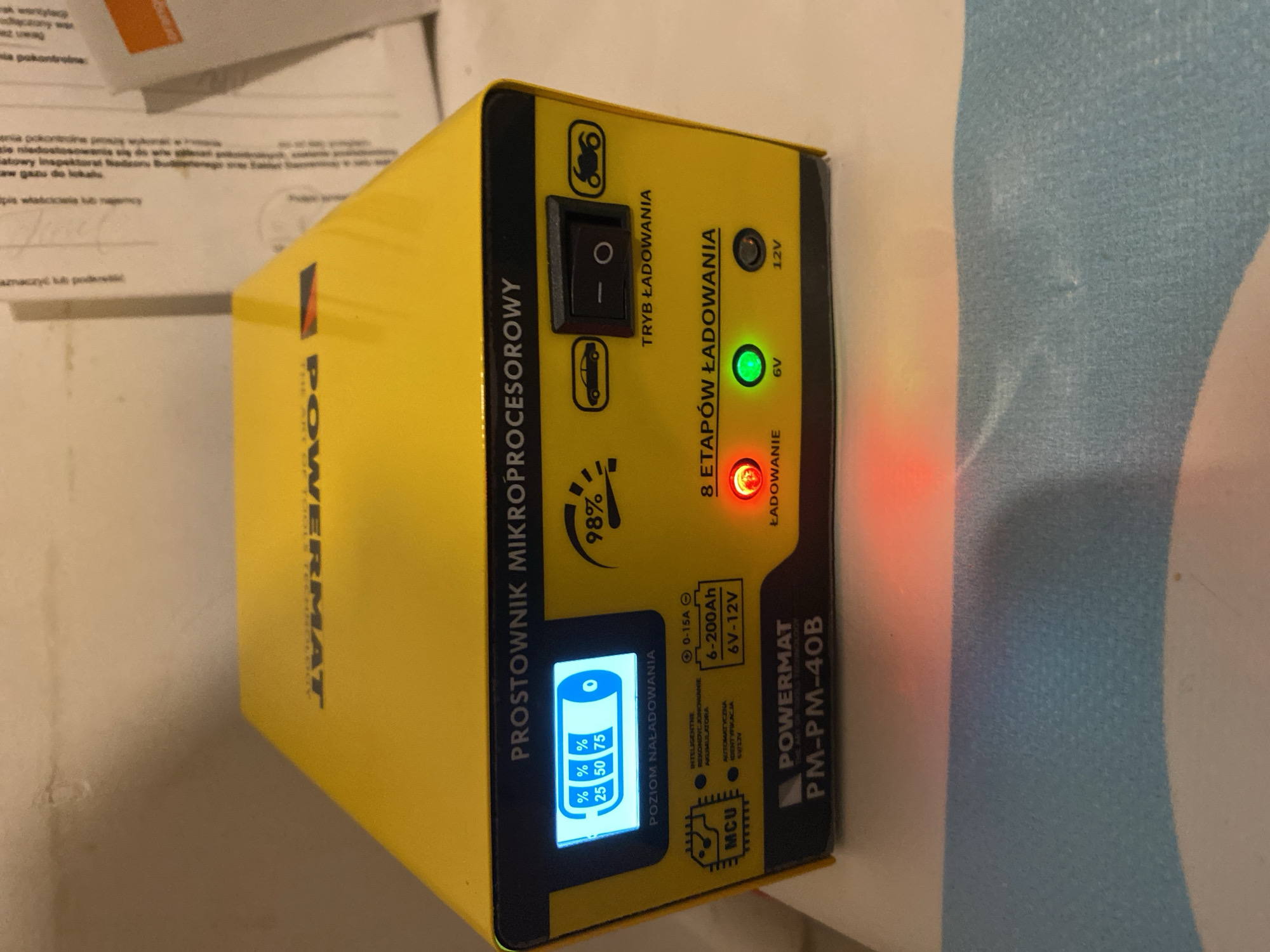 The image shows a yellow Powermat PM-PM-40B battery charger. The charger is a microprocessor-controlled device designed for charging car and motorcycle batteries. It features a digital display showing the battery charge level, with indicators for 25%, 50%, and 75% charge. There are also icons representing a car and a motorcycle, and a switch to select the charging mode. The device has LED indicators for 6V and 12V charging, with the 6V indicator currently illuminated green, suggesting it is set for 6-volt charging. A red "ŁADOWANIE" (charging) light is also visible. The charger is placed on a surface that appears to be a table or desk, covered with a light blue and white striped cloth. The background shows papers with text, some of which are in Polish, typical of documents found in an office or workshop. The lighting suggests an indoor environment. The scene does not contain any people. The image does not provide any specific information that would allow for the determination of its location within the city of Szczecin, Poland.