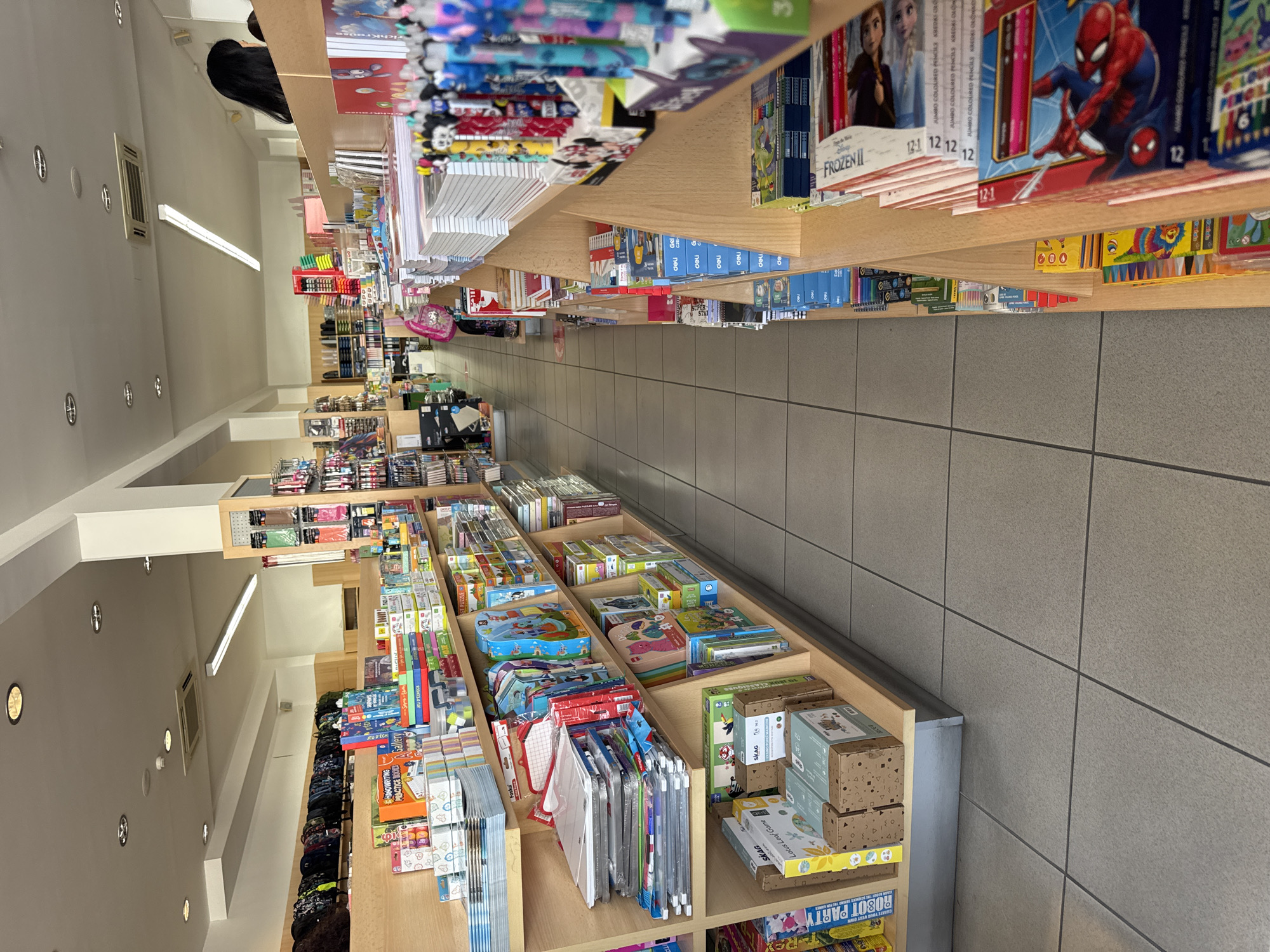 The image shows the interior of a stationery and toy store in Agios Athanasios Municipality, Cyprus. The store features aisles with shelves displaying a variety of products including backpacks, art supplies, notebooks, board games, and children's toys. In the foreground, shelves are filled with colorful items such as art sets, puzzle boxes, and stacks of notebooks. A few people are visible in the background, including a person with long black hair looking towards the right and another person partially obscured by merchandise. The store has tiled flooring and fluorescent lighting. The overall impression is of a well-stocked retail environment.