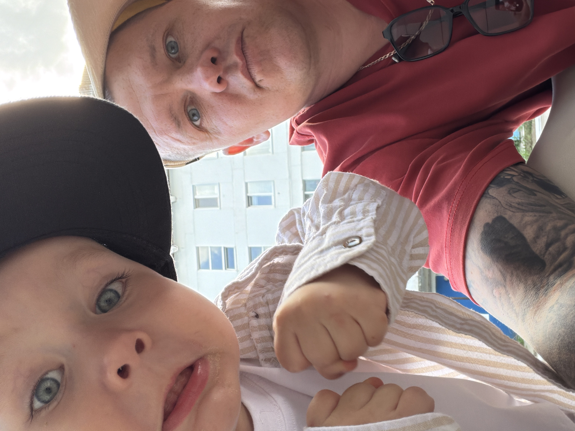In this close-up, selfie-style photograph, two individuals, an adult male and a young child, are positioned in the foreground. The child, on the left, is wearing a black baseball cap and a striped shirt. The adult male, on the right, is wearing a red hoodie and a light-colored hat. He has sunglasses hanging from a chain around his neck. Both individuals are looking towards the camera. In the background, a building with multiple windows is visible, suggesting an urban setting. The lighting indicates it is daytime, possibly outdoors given the brightness and the sky visible at the top of the frame. The image appears to have been taken on a sunny day. No specific landmarks or identifiable features place the location in Chaikovsky, Russia, but the architectural style of the building could potentially be consistent with structures found in Russian cities.