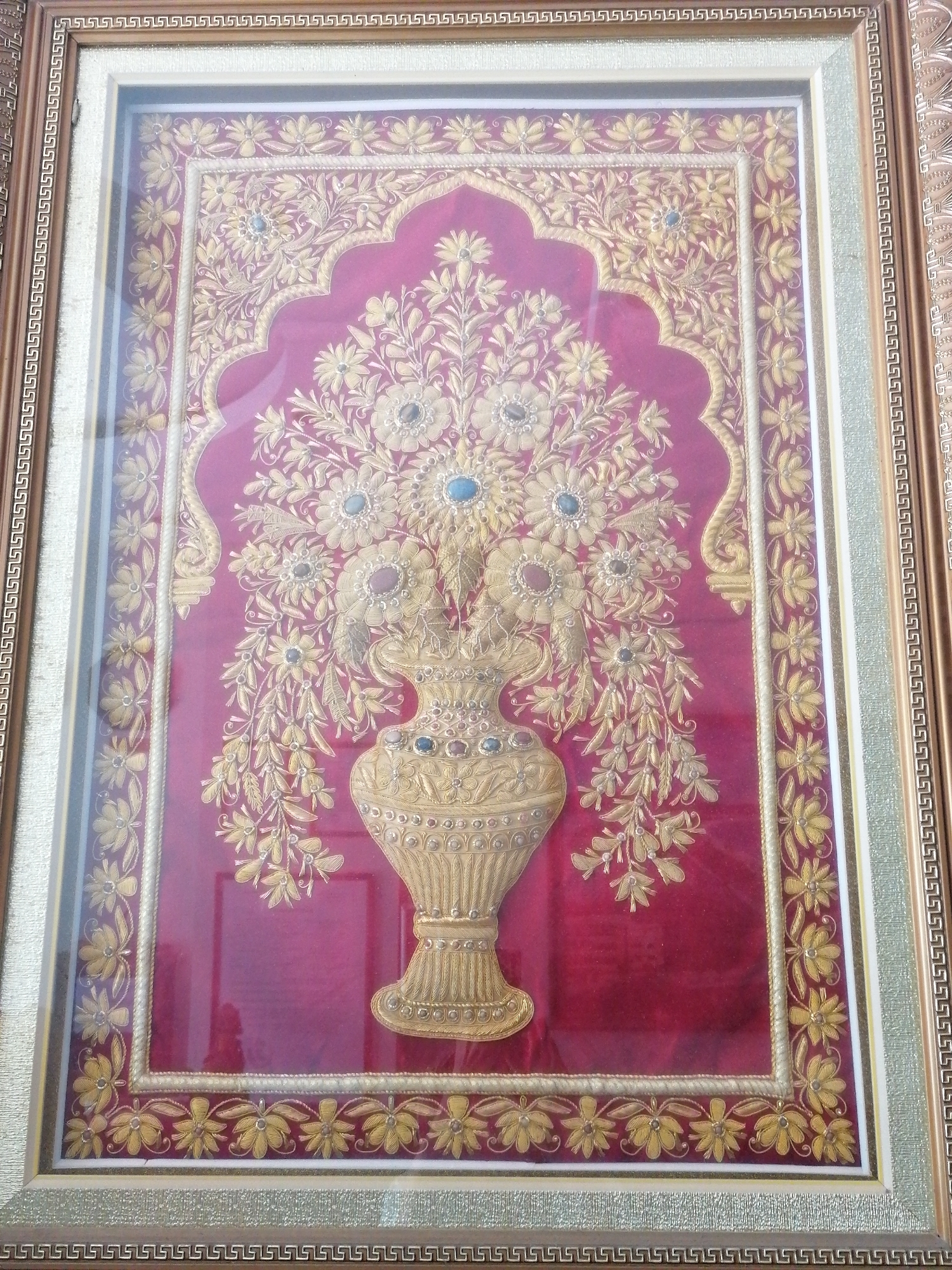 A framed textile art piece featuring a gold-colored vase filled with flowers is displayed behind glass. The background of the textile is a deep red. The art is framed with a light gold inner frame and a darker brown and gold outer frame featuring a geometric pattern. The scene appears to be indoors.