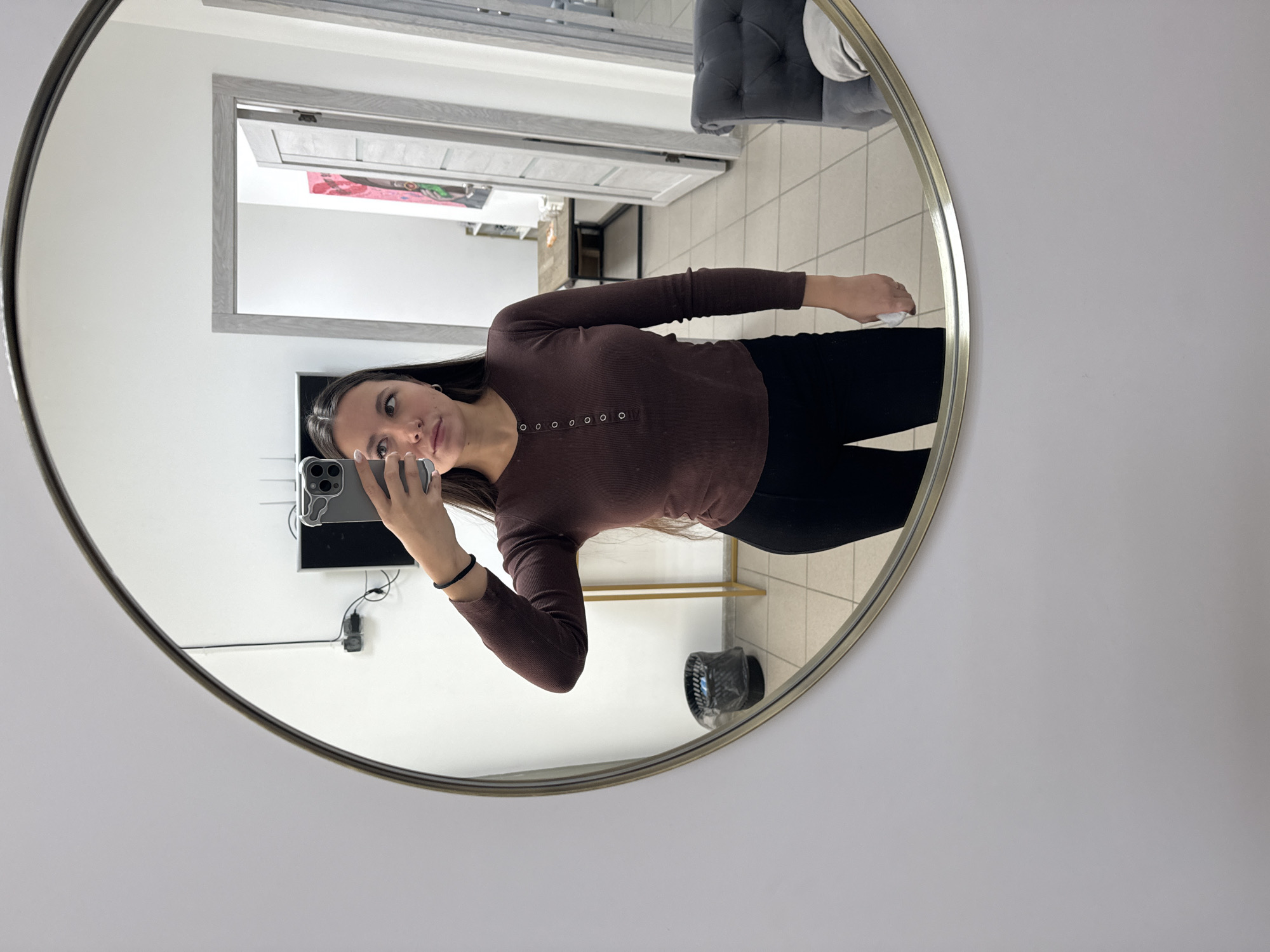 An adult female is depicted taking a mirror selfie in an indoor setting located in Odintsovo, Russia. She stands centered, reflected from the waist up in a large circular mirror with a gold-toned frame, mounted on a plain white wall. The female has long, dark hair, light-toned skin, and wears a long-sleeved, ribbed brown Henley shirt with a series of silver buttons down the front, paired with black pants. A thin black elastic band is on her left wrist, and a silver hoop earring is visible in her left ear. She holds a gray smartphone, encased in a clear or white wavy-edged cover, with her left hand, angling it towards her reflection. Her right hand is partially visible, holding a small white object, possibly a tissue or cloth. She maintains a neutral facial expression, with her lips slightly parted and eyes directed slightly upwards towards the phone.
The background, visible through the mirror's reflection, includes white walls and a light-colored tiled floor. Two doorways with light-gray frames are present. Through the left doorway, a room with a dark-colored television mounted on a white wall is visible, with cables and a black device below it. Further back in this room, a wooden table with a dark metal base and items on its surface is seen, along with two pieces of colorful artwork on the far wall (one pink/red, one green). A black waste bin with a plastic liner and a gold-toned, slender metal stand are situated to the left of the female's reflection. To the right, a gray tufted armchair or sofa section is partially visible.