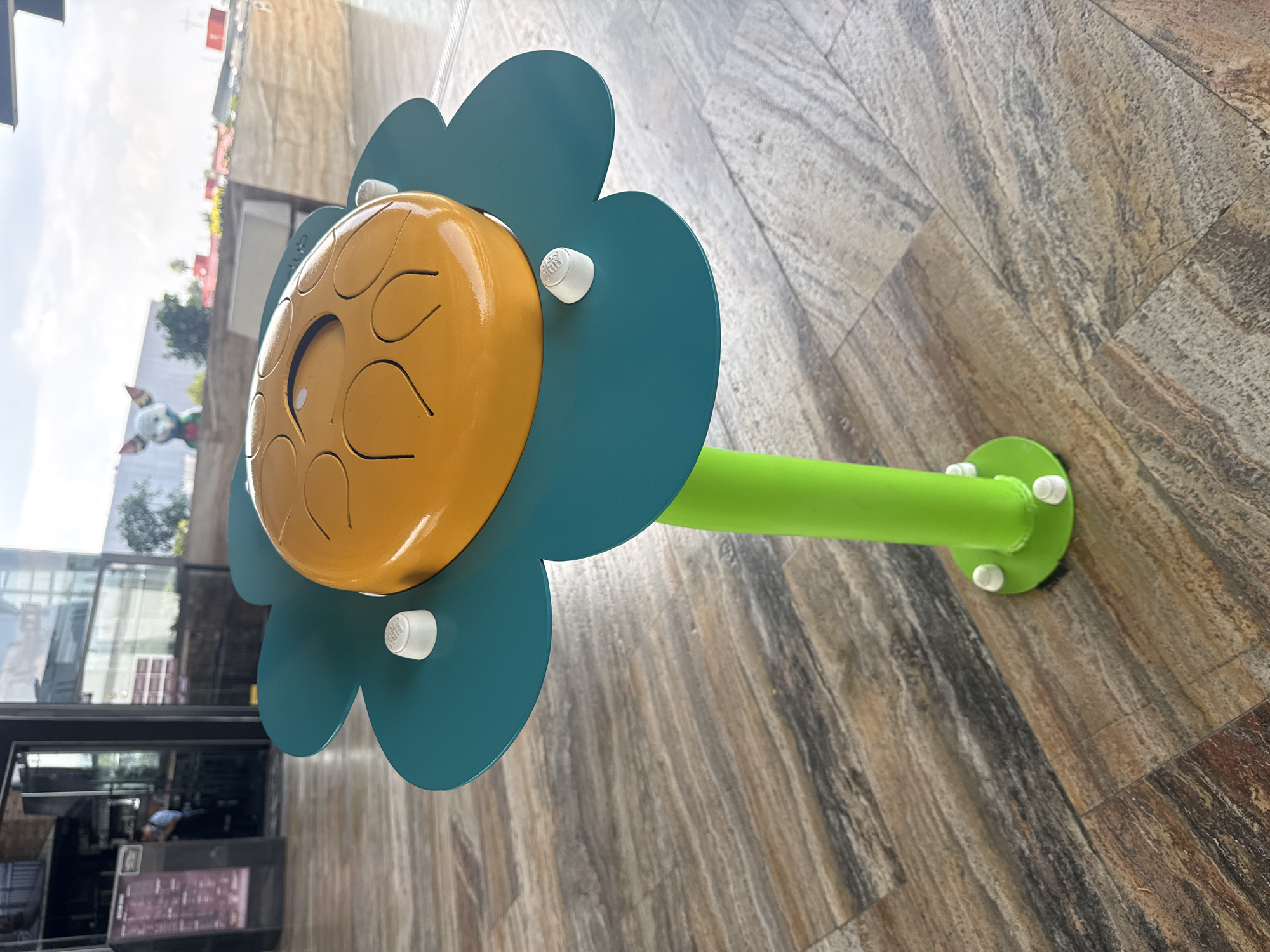 The image shows a colorful, flower-shaped musical instrument installed on a wooden tile floor, likely outdoors. The "flower" has blue petals, a yellow center with carved grooves, and white plastic caps. A green cylindrical stem supports the flower and is anchored to the floor with a round green base, also secured with white plastic caps. The instrument is located in Mexico City, Mexico, as indicated by the white caps with the words "CIUDAD DE MÉXICO" printed on them. In the blurred background, there's a building with a glass facade and a cartoon character figure. People are seen inside the building through the glass. The day seems bright, with clear visibility.