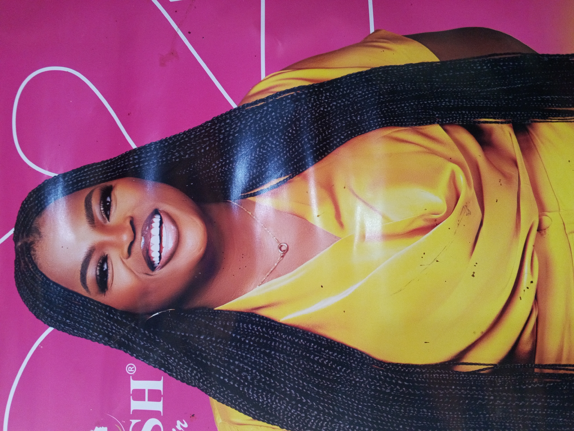 A photograph of a promotional poster featuring a woman with braided hair and a yellow outfit is situated within the city of Gwong, Nigeria. The woman is smiling, displaying her teeth, and wearing a necklace. The background of the poster is pink.