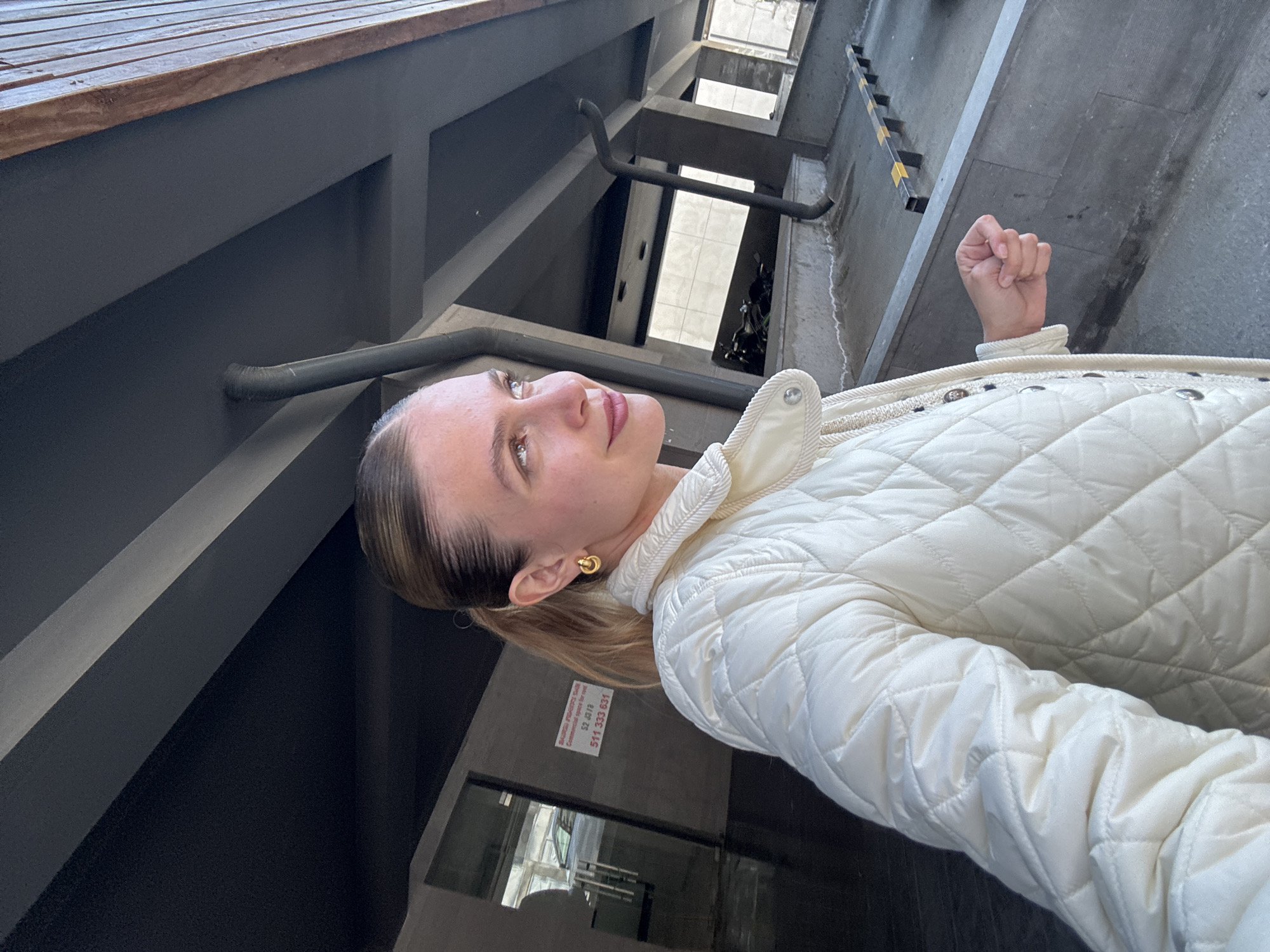 A woman is depicted from the chest up, facing away from the camera and looking upwards and to her left. She has light brown hair pulled back and wears a cream-colored quilted jacket with a high collar and a gold hoop earring on her visible ear. Her expression is neutral as she gazes upwards. Her right hand is visible, clenched into a fist, in the upper right part of the frame.
The background consists of a stark, geometric architectural setting, likely an indoor or semi-enclosed concrete structure. Dark gray walls and structural beams form sharp angles. Dark pipes or conduits are visible running along the walls. In the deeper background, multiple levels or openings can be seen, possibly part of a lightwell or an open-plan building. A metal railing with distinct yellow and black stripes is visible on the far right. A wooden ledge or surface spans the top left corner of the frame. A white sign with red text is partially visible on a wall behind the woman, displaying "SBAAN PROJECTS S.R.L. Com... space ... 2,0 / 78 ... 511 333 631". The lighting suggests it is daytime.