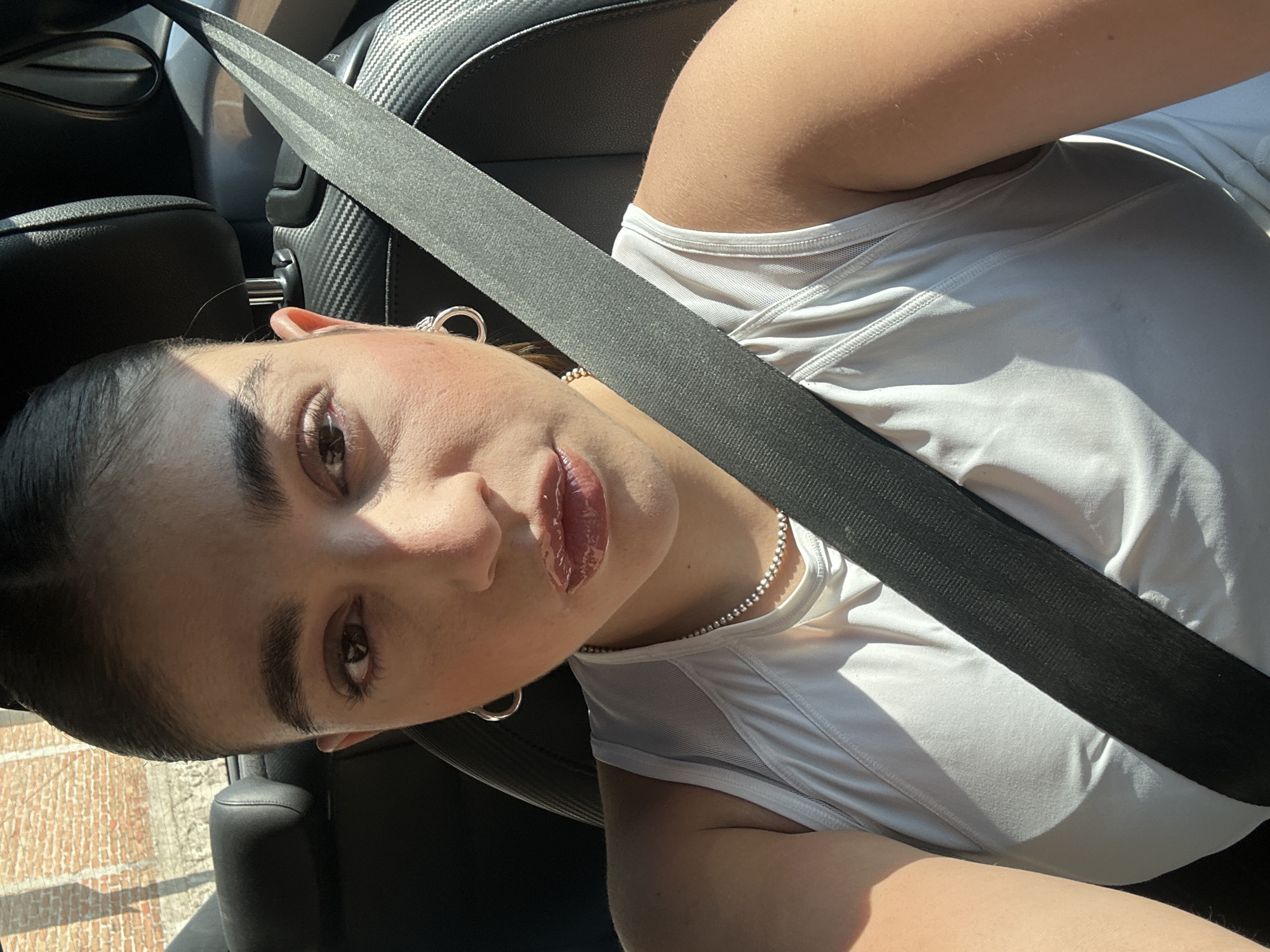A woman is sitting in a car, wearing a white tank top and a silver necklace. A seatbelt is fastened across her chest. She has brown hair pulled back, brown eyes, and is wearing lip gloss. She is looking directly at the camera. The car's interior is black leather. The location is not discernible based on the provided image. The city of San Bernardino Tlaxcalancingo, Mexico, is not verifiable based on visual elements in the image.