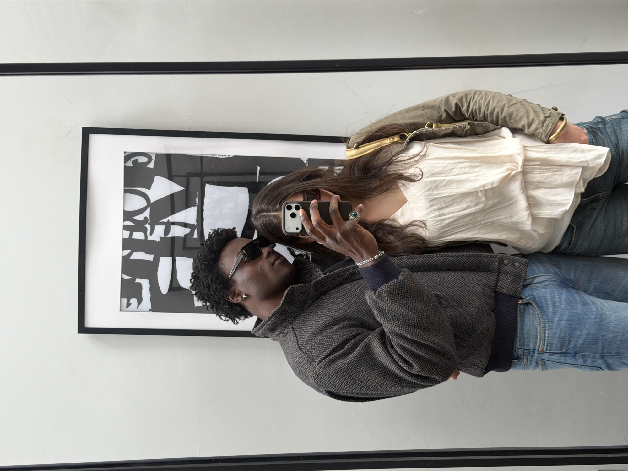 A mirror selfie captures a woman and a man in a light-colored room in London, UK. The man is on the left, wearing sunglasses and a dark jacket over a dark top and blue jeans. The woman, positioned to the right, wears a white blouse and blue jeans, and is holding the phone taking the photograph. A framed black and white graphic print hangs on the wall behind them. The mirror's black frame is visible at the top and bottom of the image.