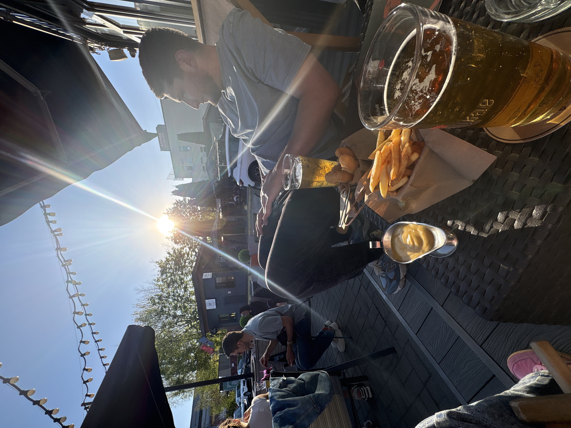 Here is a description of the image:
The image shows an outdoor dining scene, likely at a restaurant or bar in Minsk, Belarus. A man in a grey shirt sits at a table with a beer and a paper container of french fries and fried dumplings, served with a side of sauce. Another person, out of focus, is kneeling on the ground nearby. In the background, there is a street scene with buildings, trees, and people. String lights are strung overhead. The sun is bright, creating lens flare and casting shadows.