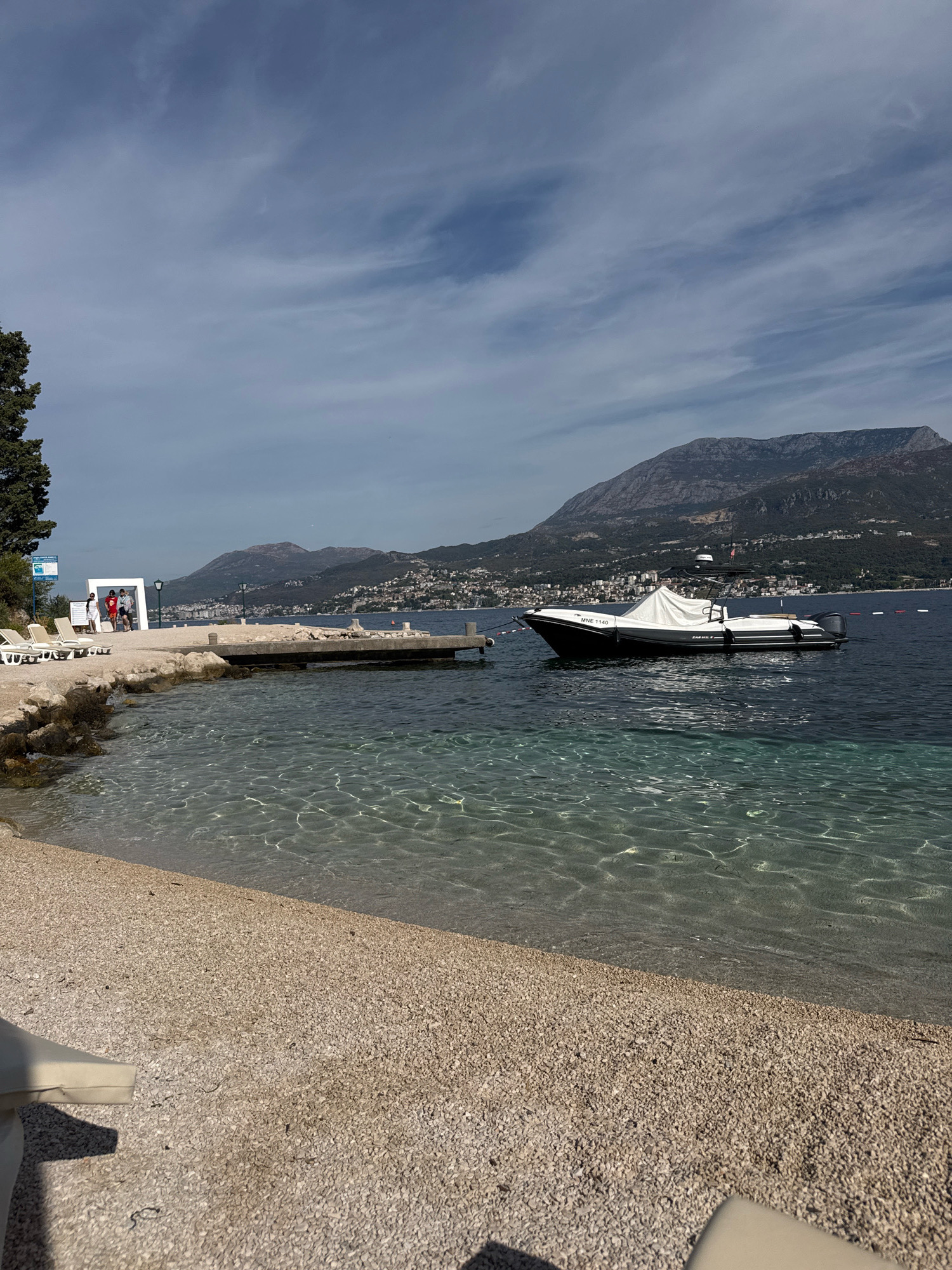 The image depicts a scenic view of a coastline on a clear day, with a body of water stretching towards distant mountains. In the foreground, a pebble beach leads to the calm, transparent water, where sunlight creates shimmering patterns on the sandy seabed. A white lounge chair is partially visible in the lower left corner.
Further out on the water, a sleek, dark-hulled boat with a white cover is moored. The boat has the registration "MNE 1140" visible on its side. Adjacent to the boat, a concrete pier extends into the water, with a few people standing near its end. One person is wearing a red shirt, another a white shirt, and the third is partially obscured. A white modern art-like structure stands behind them.
A lush green tree is situated to the left of the pier, and beyond that, a signpost is visible. The background is dominated by a range of imposing mountains, dotted with buildings that suggest a town or city nestled along the coastline. The sky is a bright blue with wispy white clouds. The overall scene conveys a sense of tranquility and a picturesque coastal environment.