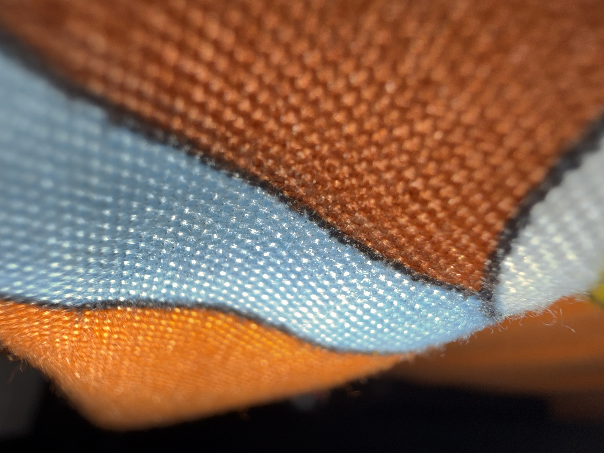 The image is a close-up, macro photograph of a fabric surface. The fabric appears to be woven and has a textured appearance with visible threads. There are at least three distinct color bands: a deep orange on the bottom and top right, a light blue in the middle, and a white band along the left edge, bordered by a thin black line. The light blue band has a shimmering or slightly reflective quality, possibly due to the way light interacts with its weave. Small, wispy fibers are visible along the edges of the fabric, particularly where the white and blue sections meet. The lighting creates highlights and shadows, emphasizing the texture of the material. The background is dark and out of focus. There are no people, animals, or recognizable city elements; the image focuses exclusively on the material's texture and color. The location in Uyo, Nigeria, cannot be determined from the visual information presented in the image.