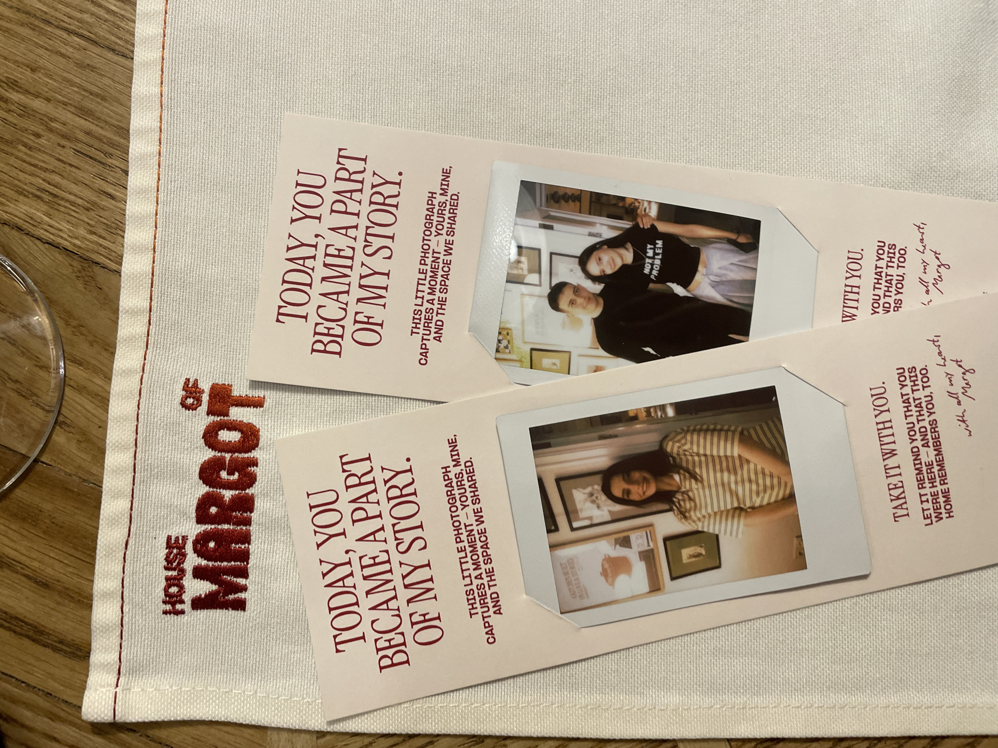 The image shows a section of a dining table at a location named "House of Margot". A white cloth napkin with red embroidered lettering and a red hemmed edge sits on the table. On top of the napkin are two identical cards. Each card has the same text in red: "TODAY, YOU BECAME A PART OF MY STORY." and "THIS LITTLE PHOTOGRAPH CAPTURES A MOMENT - YOURS, MINE, AND THE SPACE WE SHARED." Attached to each card is a Polaroid photograph. One Polaroid shows a woman wearing a black t-shirt that says "NOT MY PROBLEM" standing with a man. The other Polaroid shows a woman in a striped shirt. The cards also have the text "TAKE IT WITH YOU. LET IT REMIND YOU THAT YOU WERE HERE - AND THAT THIS HOME REMEMBERS YOU, TOO. with all my heart, Margot" and "WITH YOU. YOU THAT YOU ND THAT THIS RS YOU, TOO. In all my heart, Margot". The location is in Yerevan, Armenia. A glass is visible in the corner of the image.