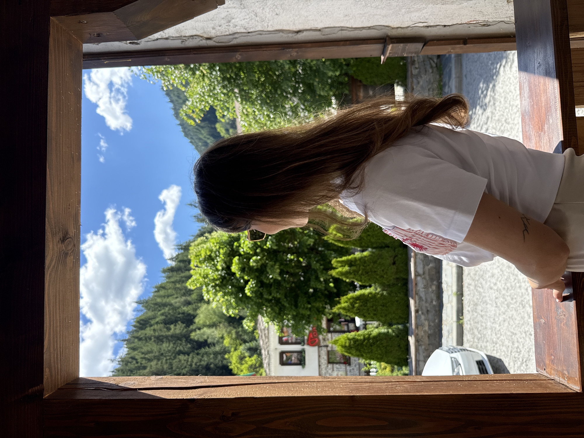 A medium shot shows a woman with long brown hair, wearing sunglasses and a white t-shirt, standing and looking out from a wooden window frame. The background reveals a scenic view of Shiroka Laka, Bulgaria, with a cluster of traditional white houses nestled among lush green trees and a steep, forested mountainside under a bright blue sky with scattered white clouds. In the foreground, a white car is partially visible parked on a cobblestone street. The woman has a tattoo on her left forearm.