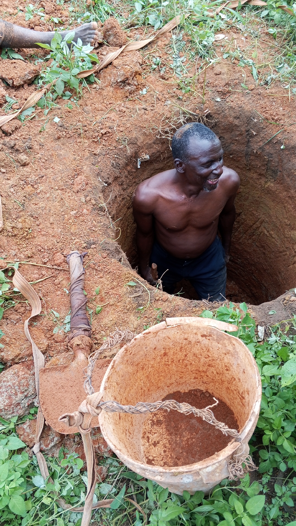 The image shows a bare-chested, dark-skinned man standing inside a roughly dug, circular hole in the ground. He is wearing dark pants. A shovel and a bucket containing soil are on the ground near the hole's edge. The surrounding area is covered with reddish-brown soil and patches of green vegetation. The man appears to be in the process of digging or has recently finished. A foot can be seen in the background. The man has a smile. The location is identified as Bukuru, Nigeria.