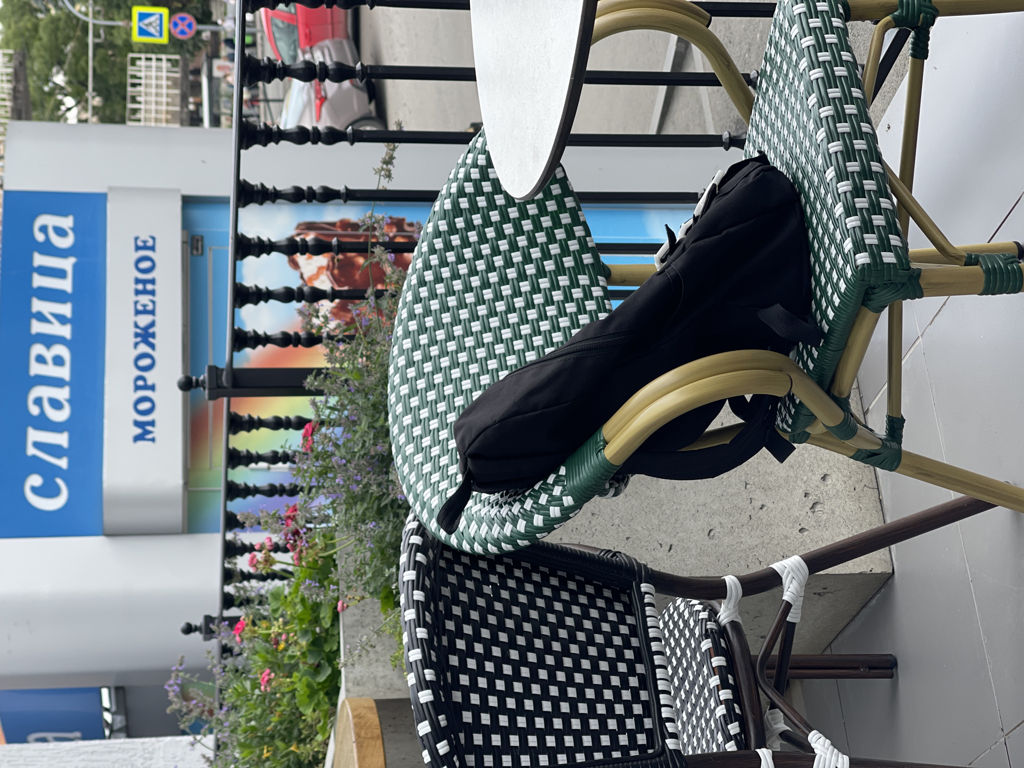 The image shows a close-up of two outdoor cafe chairs and a table, with a black backpack draped over the back of one chair. The chairs have a woven pattern of green and white on the seats and backs, and the frames appear to be made of wood or a similar material. The chair on the left has a black and white woven pattern. In the background, there is a building with signage in Russian that reads "СЛАВИЦА" and "МОРОЖЕНОЕ," indicating an ice cream shop called "Slavitsa." There is also a black metal fence with decorative posts, and some flowering plants. Beyond the fence, cars and traffic lights are visible, suggesting the cafe is located in an urban area. The setting is an outdoor seating area of a cafe, likely in Izhevsk, Russia, given the Russian language signage.