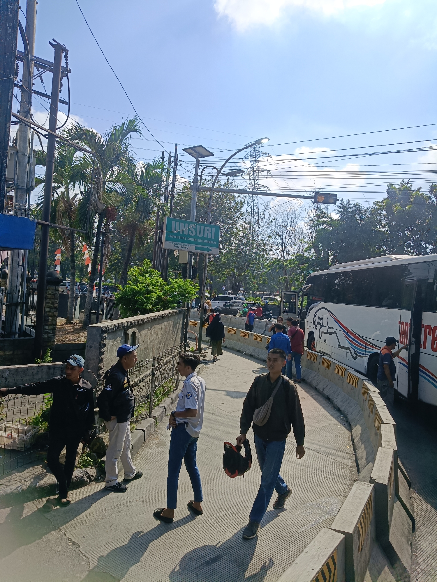 The image captures a bright, sunny day in what appears to be an urban or semi-urban area. A large white bus with a black and red logo depicting a running animal is parked on the right side of the road. A traffic light is visible in the upper right corner. In the foreground, several individuals are present on a paved pathway that curves to the left, separated from the road by concrete barriers with yellow markings.
On the pathway, a man in a dark jacket and blue jeans, carrying a helmet, walks towards the viewer. To his left, another man in a blue shirt and dark pants stands with his hands in his pockets. Further left, a man in a black jacket and dark pants leans against a metal fence, looking towards the left edge of the frame. Two other men in casual attire are conversing near him.
On the left side of the image, a street sign with green lettering that reads "UNSURI" and below it "UNIVERSITAS SUNAN GIRI SURABAYA" is mounted on a pole. Palm trees and other greenery are visible behind the sign. Power lines and utility poles are prominent in the background, and a tall transmission tower can be seen in the distance. The overall scene suggests an active street with pedestrians and vehicular traffic. The location is identified as Kedungrejo, Indonesia.
